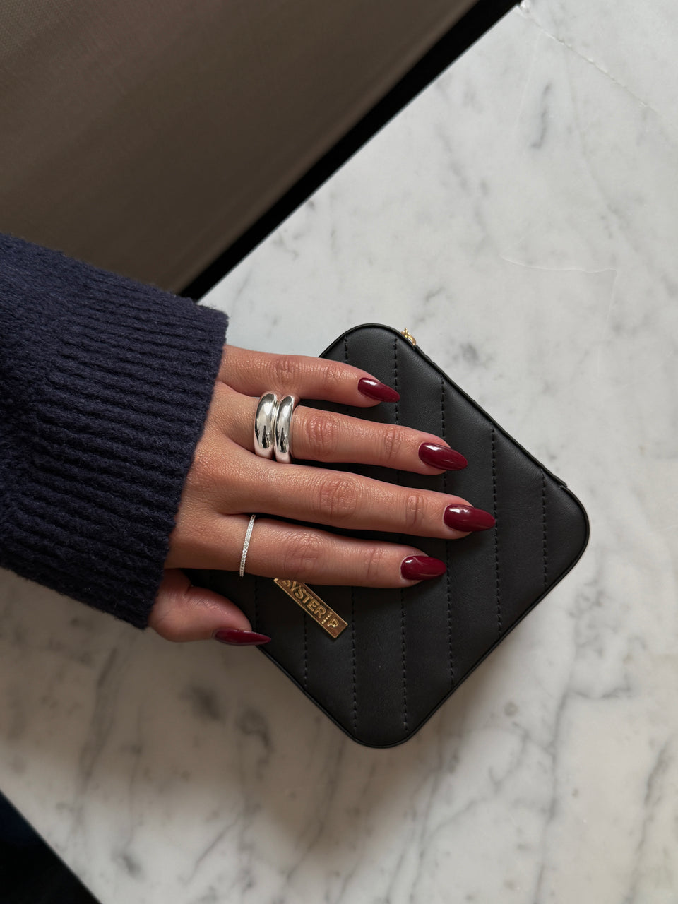 A model's hand, wearing a bold double-band silver ring and a thin sparkling pavé band, rests upon the small black quilted Syster P jewelry case.