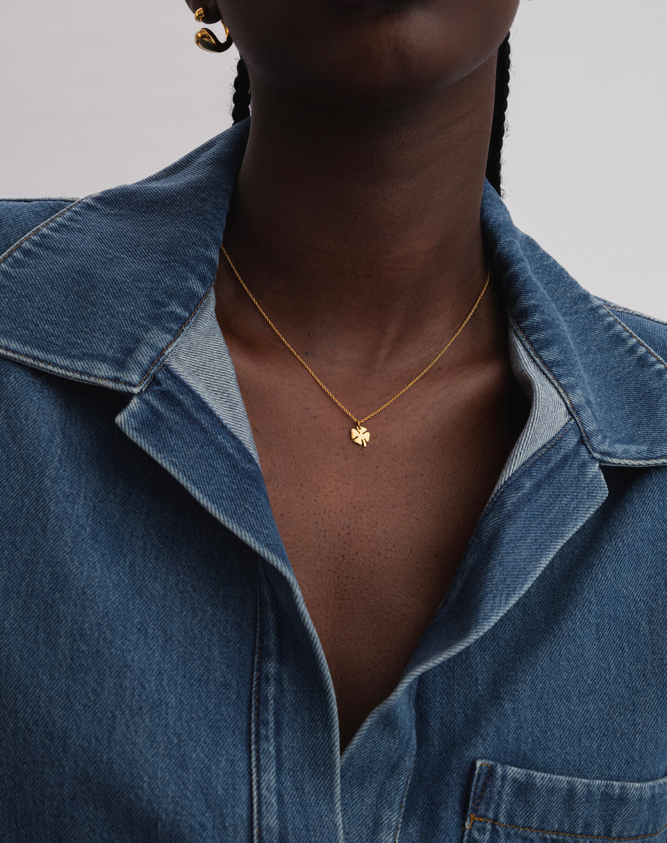 A close-up shot of a model wearing the Bring Me Luck Necklace in gold, featuring a delicate chain and a small four-leaf clover pendant. A gold earring is also visible.
