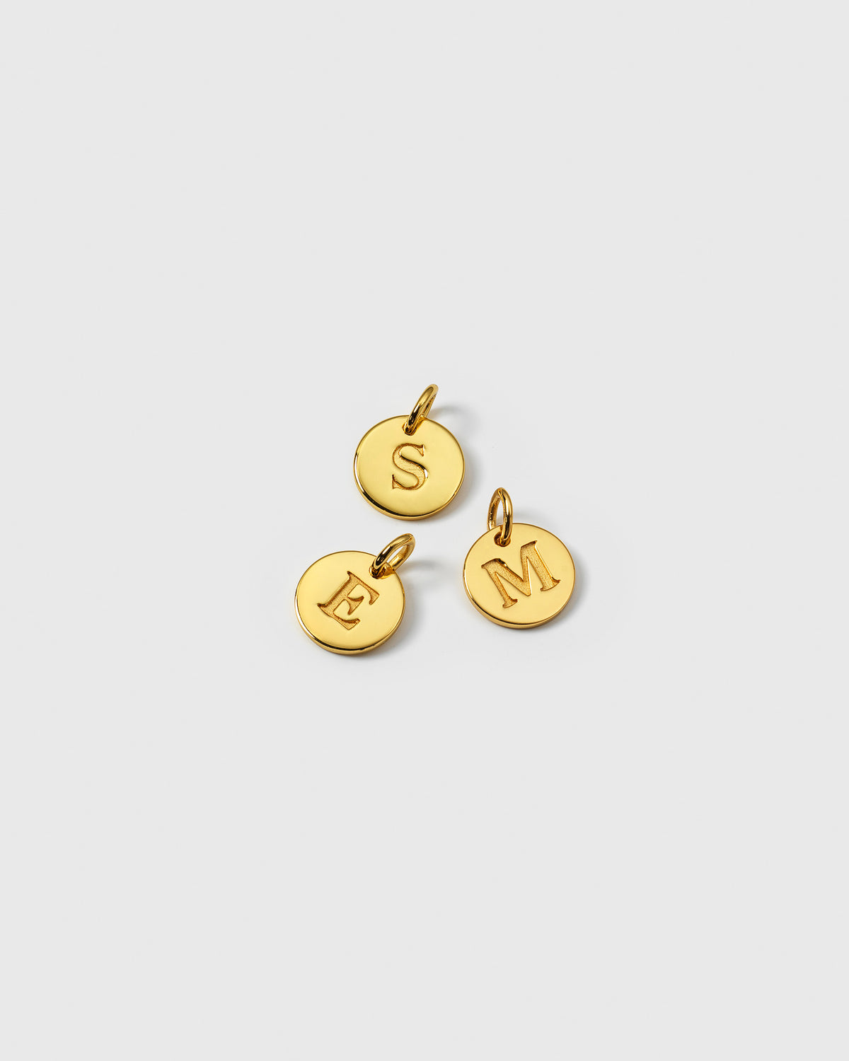 A product shot of three Beloved Mini Letter Gold pendants, showcasing the small, circular charms with the letters S, E, and M engraved on them.