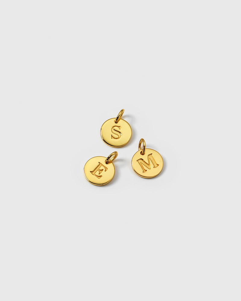 A product shot of three Beloved Mini Letter Gold pendants, showcasing the small, circular charms with the letters S, E, and M engraved on them.