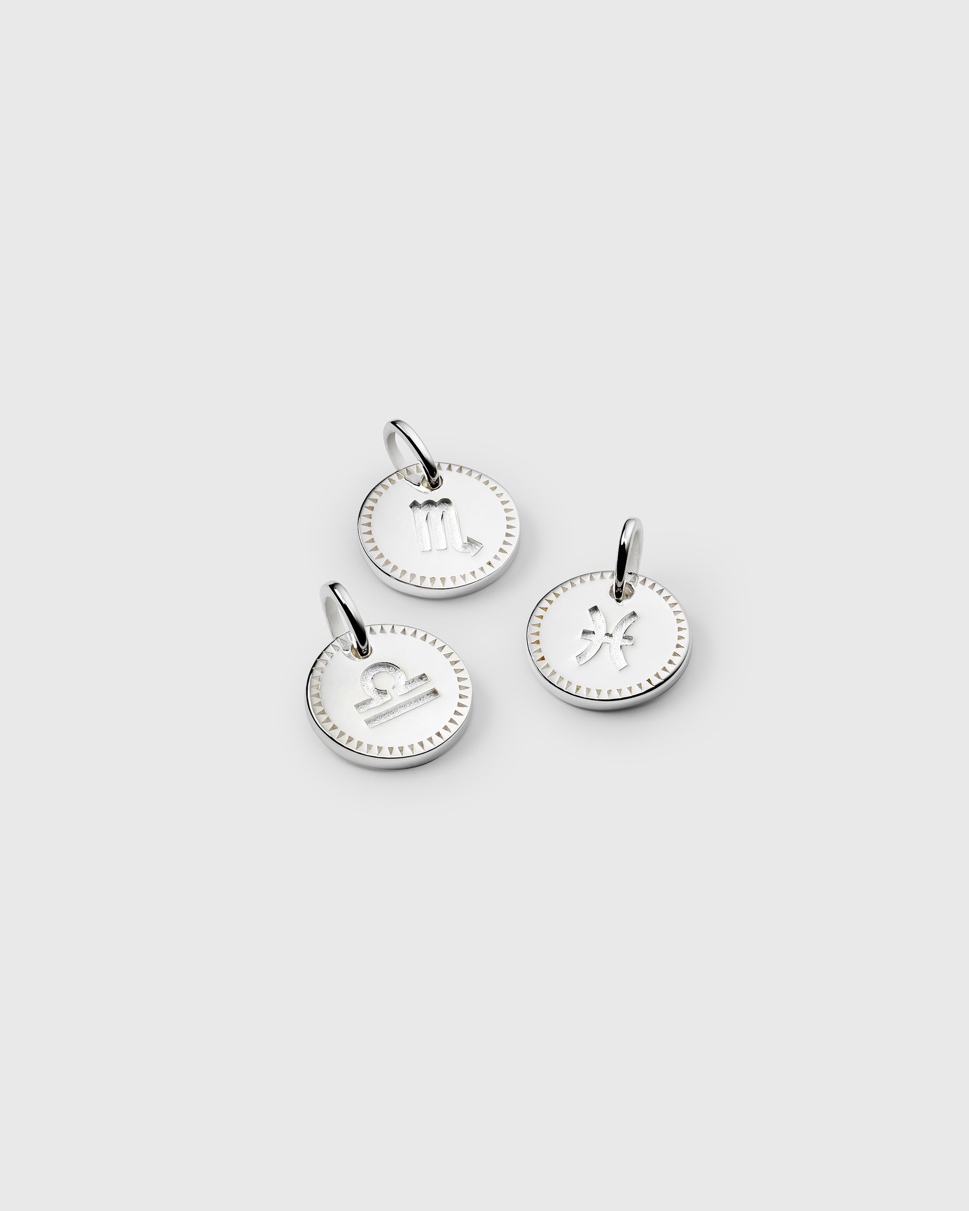A product shot of three Beloved Zodie Pendants in silver, displaying the engraved zodiac signs for Scorpio, Libra, and Pisces within a decorative border.