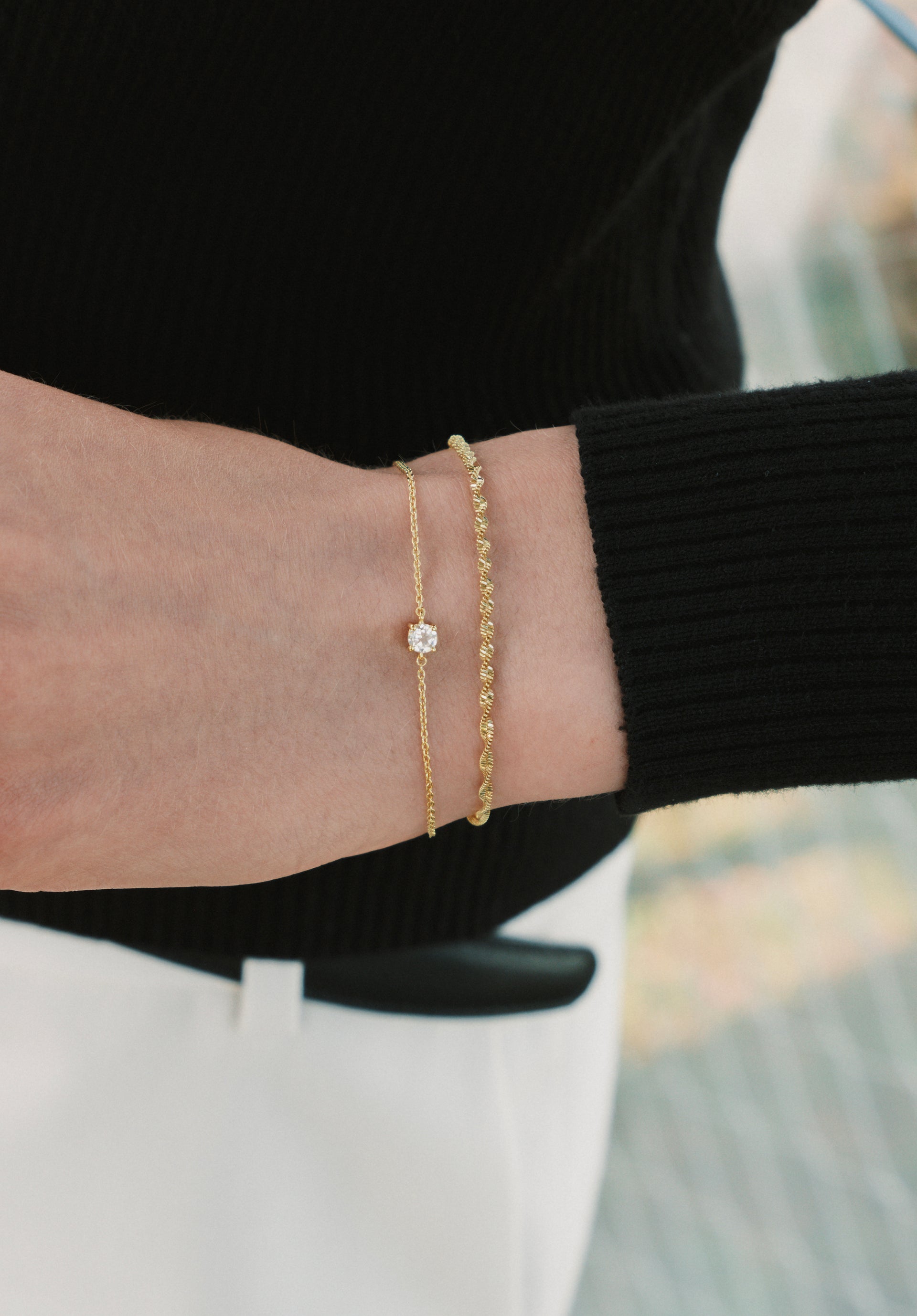 A close-up of a model's wrist wearing the Herringbone Twisted & Solo Nova Bracelet Set in gold. The set features a twisted herringbone chain bracelet stacked with a delicate chain bracelet with a single clear stone.