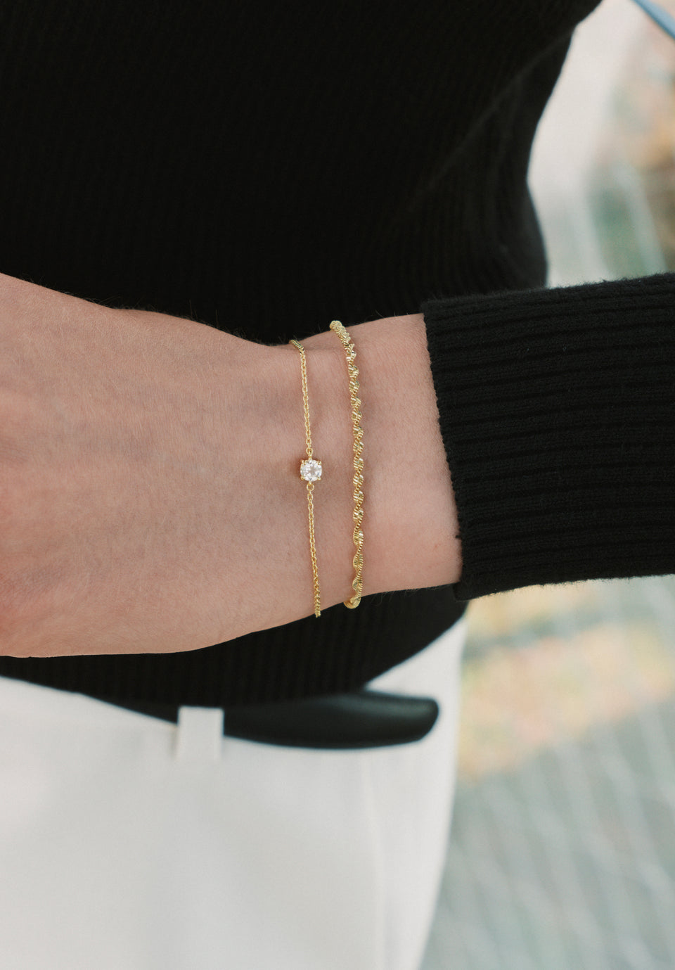 A close-up of a model's wrist wearing the Herringbone Twisted & Solo Nova Bracelet Set in gold. The set features a twisted herringbone chain bracelet stacked with a delicate chain bracelet with a single clear stone.