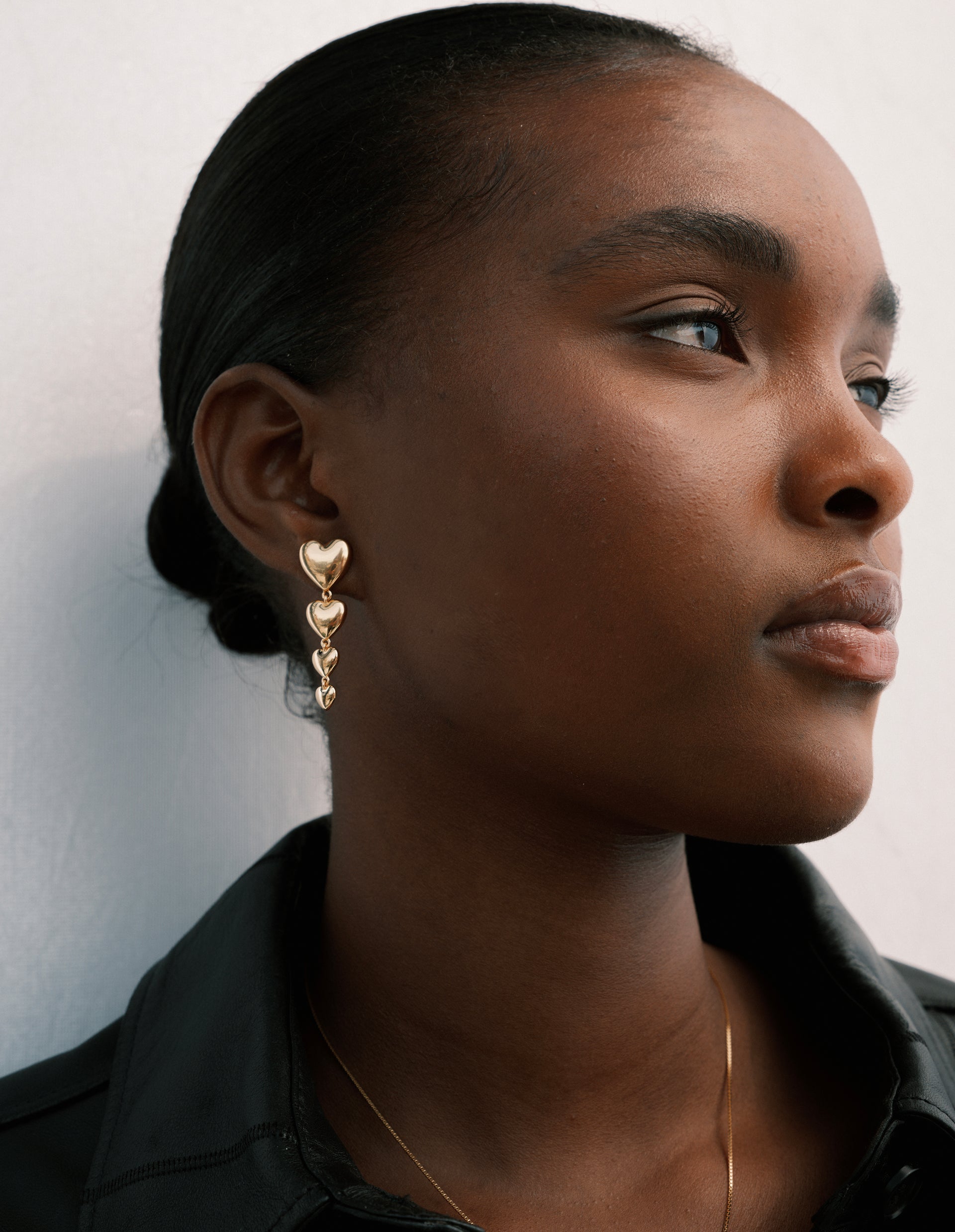Close-up of a model's profile showing the Darling Earrings in gold, which feature four polished hearts in a dangling design, along with a fine gold necklace.