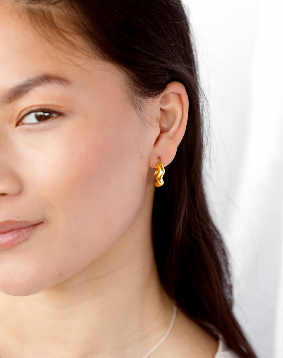 A close-up shot of a model wearing the Bolded Wavy Earrings in gold, highlighting their shiny and wavy design. A delicate silver chain necklace is also visible.
