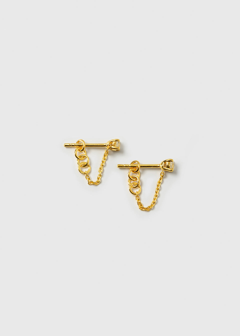 A product shot of the Minimalistica Solo Nova Earrings in gold, showcasing the pair of studs each featuring a white topaz and a delicate, draping chain.