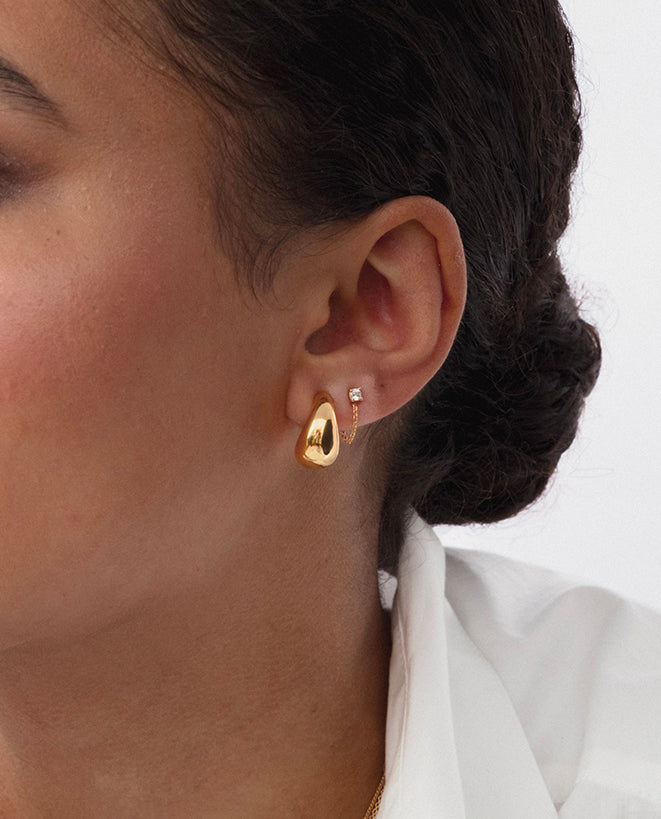 A close-up of a model wearing the gold Minimalistica Solo Nova Earrings, showcasing the design with a small huggie hoop and a delicate chain attached to a white topaz stud.