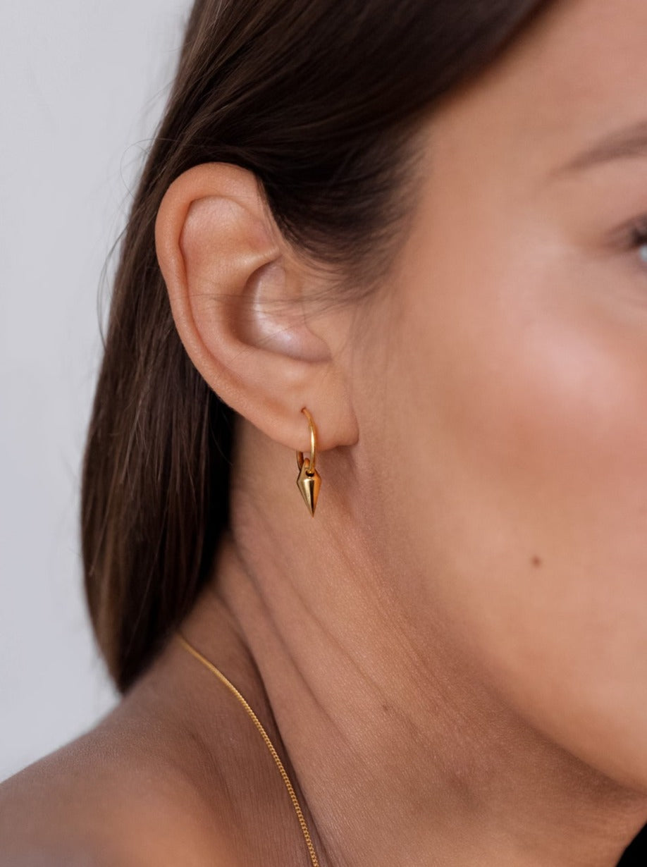 A close-up of a model wearing the Harvey Small Spike Hoops in gold, featuring a small hoop with a dangling spike charm, and a thin gold chain necklace.