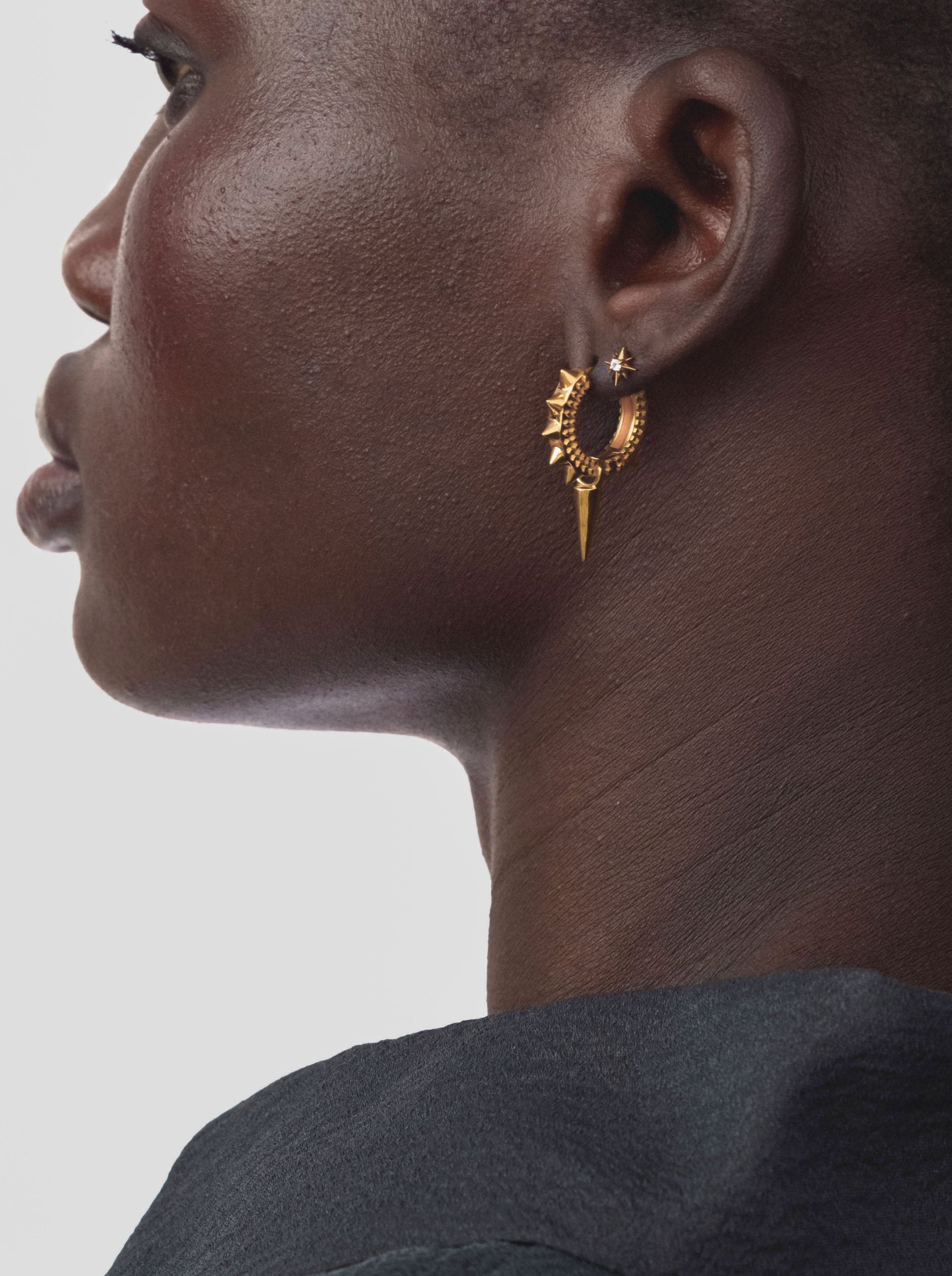 A close-up of a model wearing the Harvey Heavy Spike Hoops in gold, which feature a textured surface with multiple small spikes and a larger dangling spike, alongside a small star-shaped stud in her upper ear.