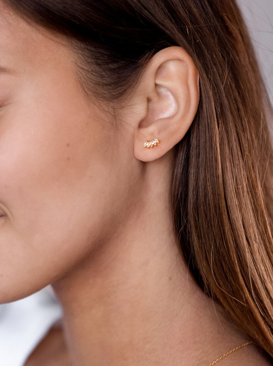 A close-up shot of a model wearing the Theodora Earrings in gold, featuring a delicate, curved design adorned with sparkling cubic zirconia stones.
