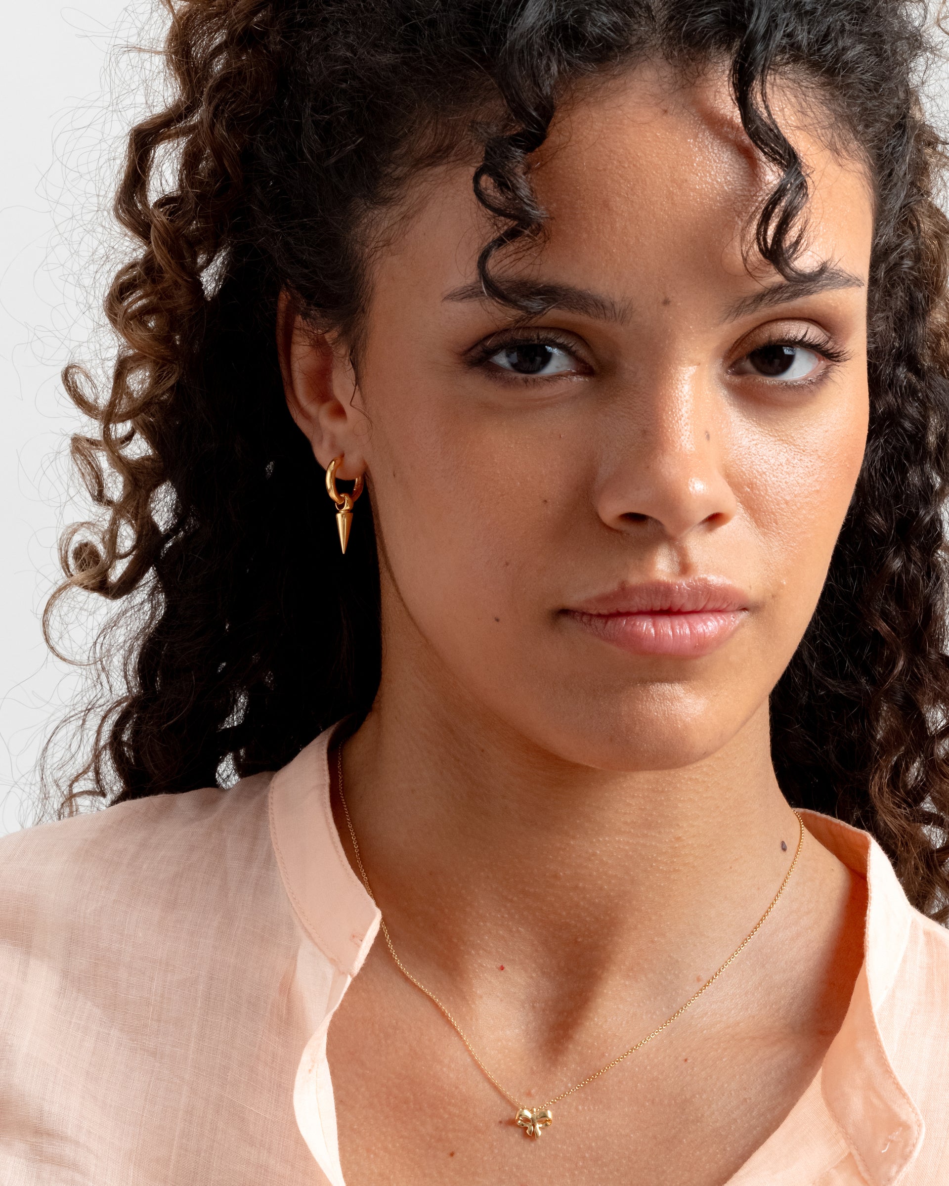 A close-up of a model wearing the gold Juno Hoop with a dangling spike charm, along with a fine gold necklace featuring a bow pendant.