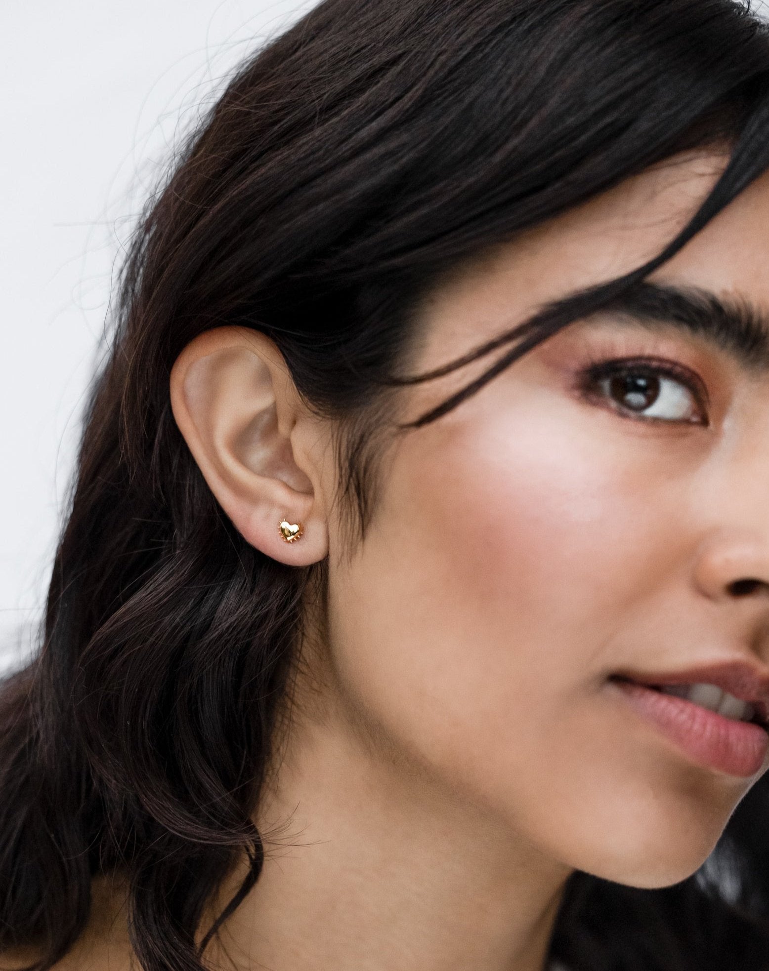 A close-up image of a model wearing the True Love Spike Heart Studs in gold, showing the small heart-shaped earring with spike details on her earlobe.