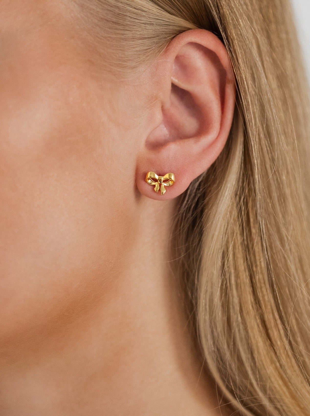 A close-up of a model wearing the Rosie Mini Studs in gold, a small stud earring shaped like a delicate bow.