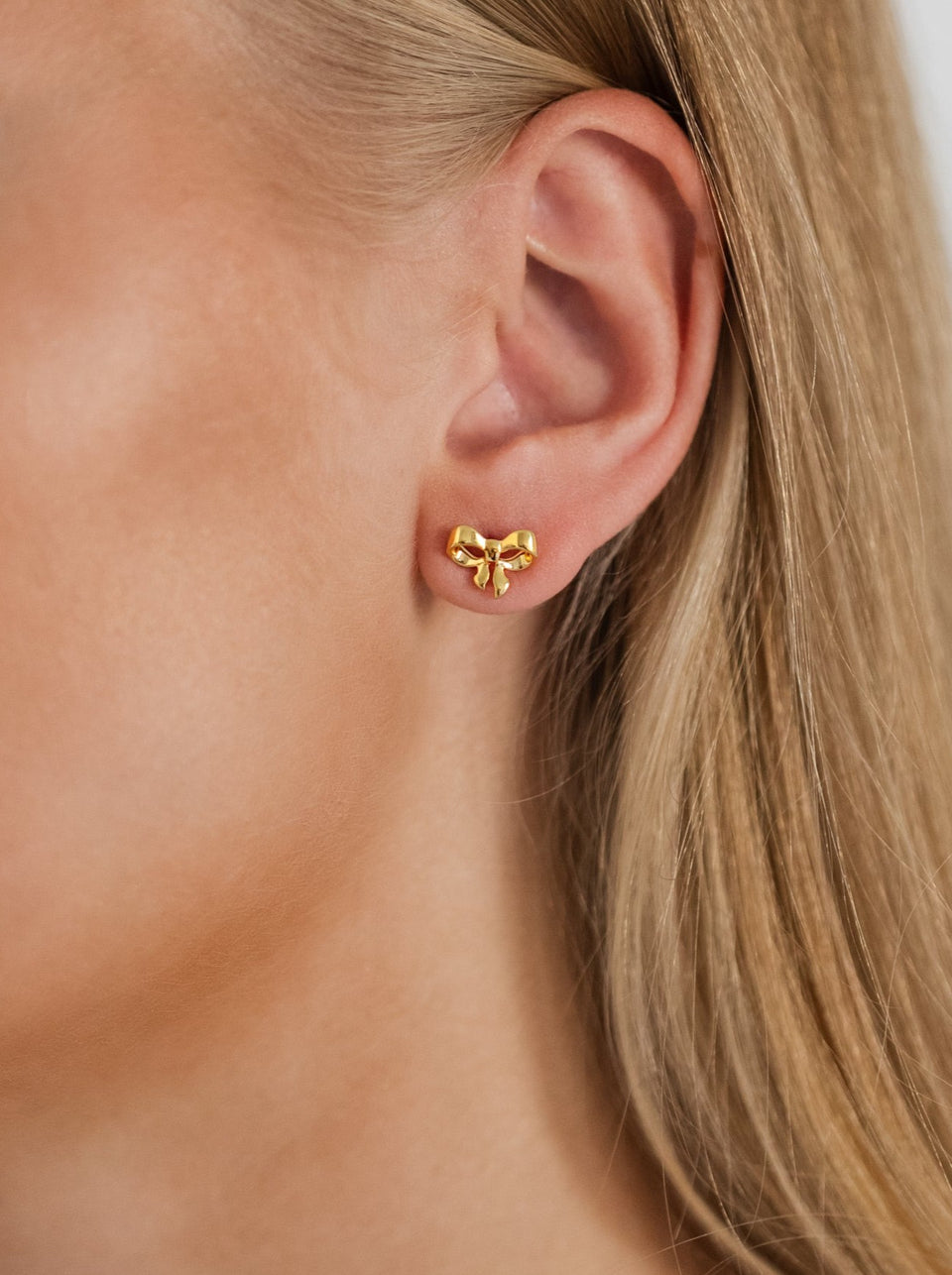 A close-up of a model wearing the Rosie Mini Studs in gold, a small stud earring shaped like a delicate bow.