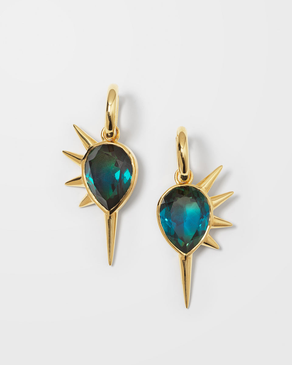 A product shot of the Queen Of Spikes Earrings in gold, featuring a pair of hoops with dangling pendants that have a large teal quartz stone surrounded by gold spikes.