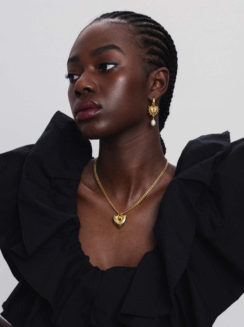 A model styles the gold Fat Heart Necklace, featuring a chunky chain and a large heart pendant, with a Drama Queen Heart Pearl Earring that has a spiked heart charm and a pearl drop.