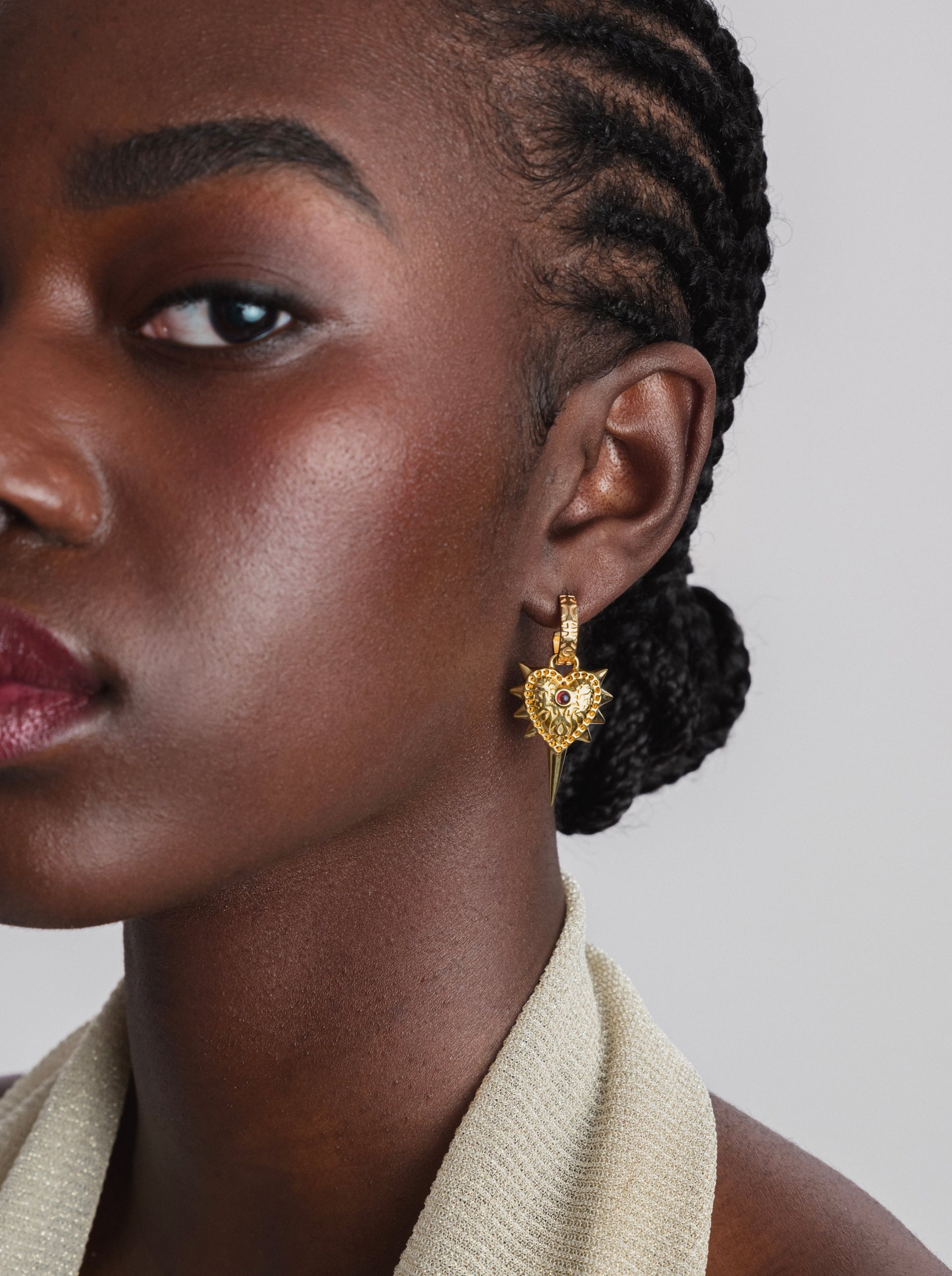 A close-up shot of a model wearing the Drama Queen Heart Spike Earring in gold, which features an intricate heart-shaped pendant with spikes and a red stone.