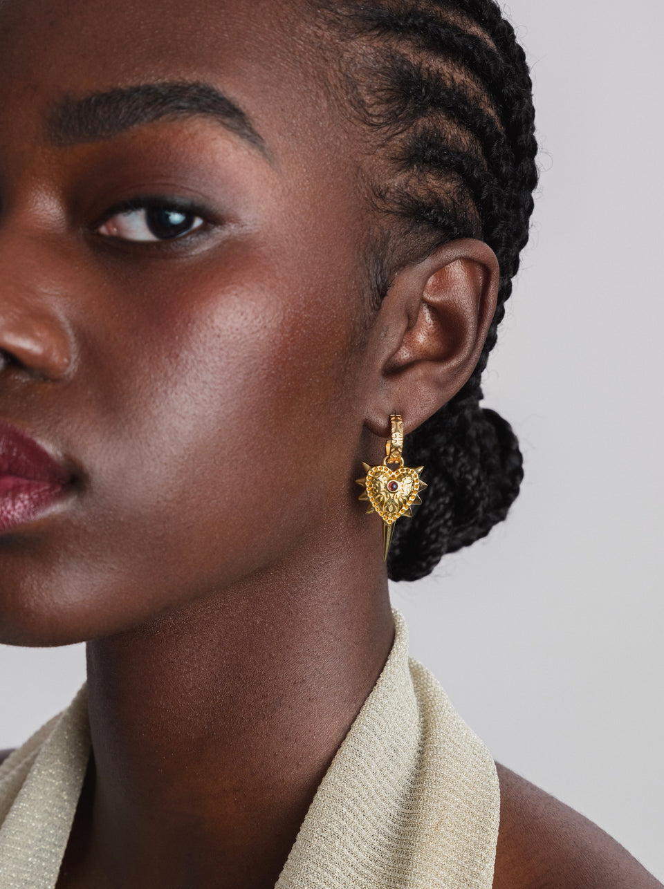 A close-up shot of a model wearing the Drama Queen Heart Spike Earring in gold, which features an intricate heart-shaped pendant with spikes and a red stone.