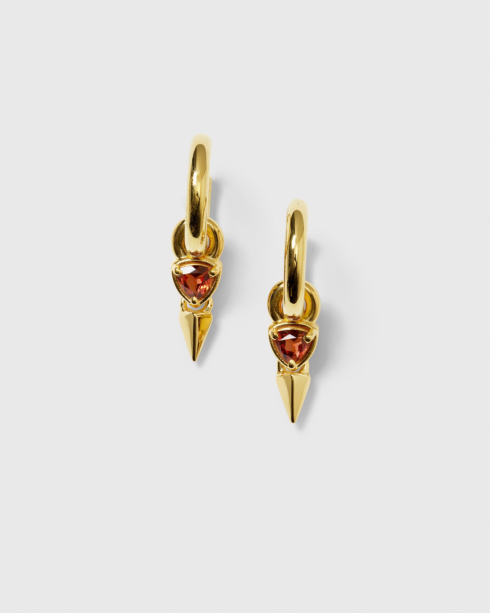 A product shot of the Mrs H Spike Earrings in gold, featuring a chunky hoop with a dangling charm composed of a triangular garnet and a pointed spike.