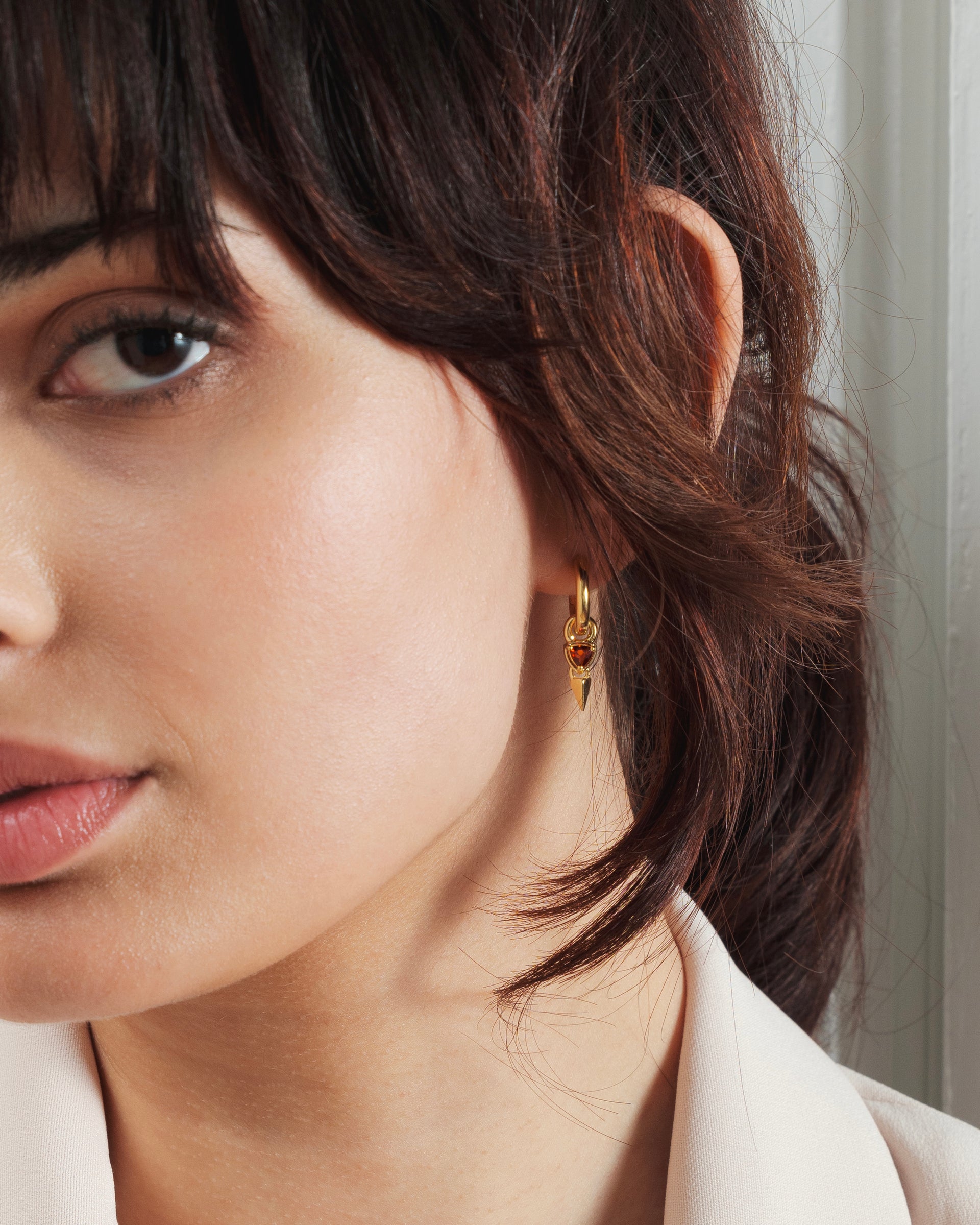 A close-up shot of a model wearing the Mrs H Spike Earrings in gold. The earring consists of a gold hoop from which a red garnet stone and a small spike dangle.