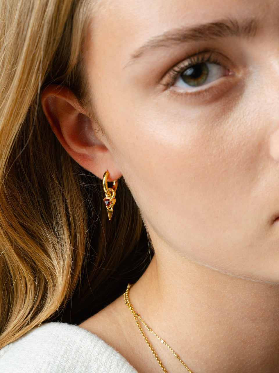 A close-up of a model wearing the Mrs H Spike Earrings in gold, featuring a hoop with a dangling red garnet and spike charm, styled with a delicate gold necklace.
