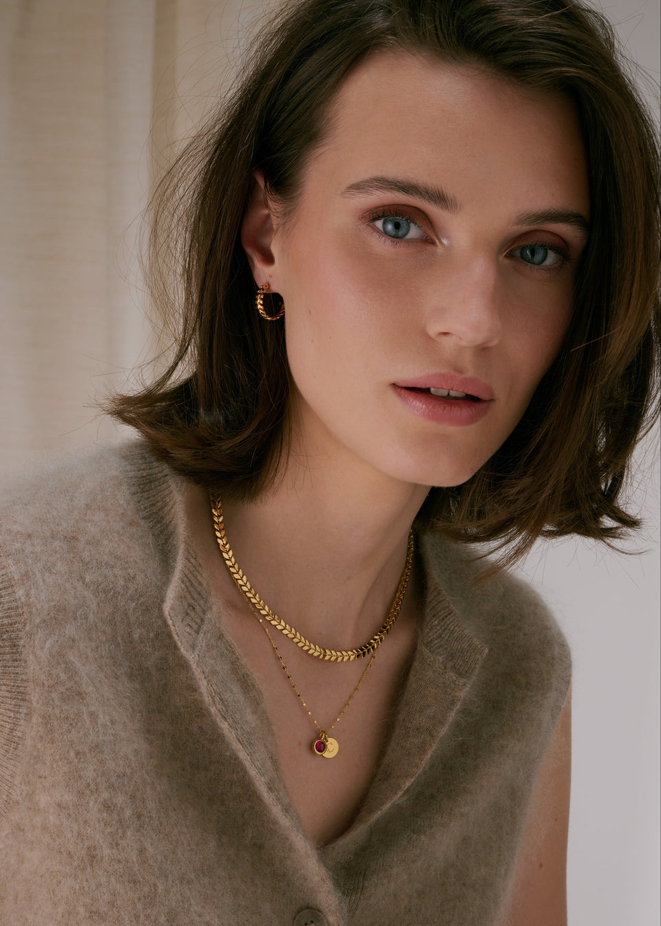 Gold hoop earrings with a leaf-like pattern, paired with two layered gold necklaces‚Äîone thick chain with a leaf pattern, and one fine chain with a pendant featuring a ruby, July's birthstone, and an initial charm.
