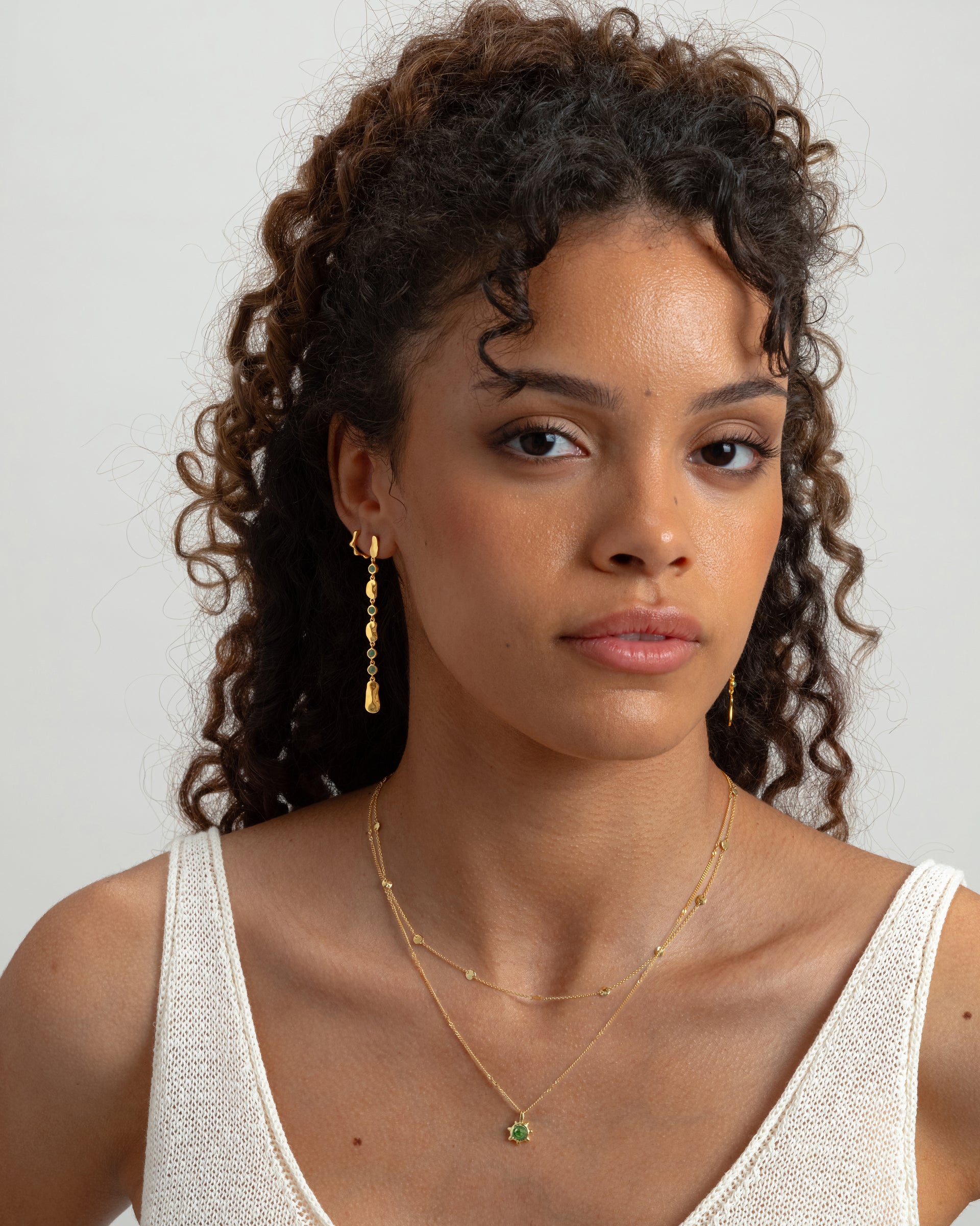 A model styled with Syster P jewelry, wearing two layered gold necklaces‚Äîone a simple chain with small discs and the other with a green gemstone pendant‚Äîand two different dangling gold earrings.