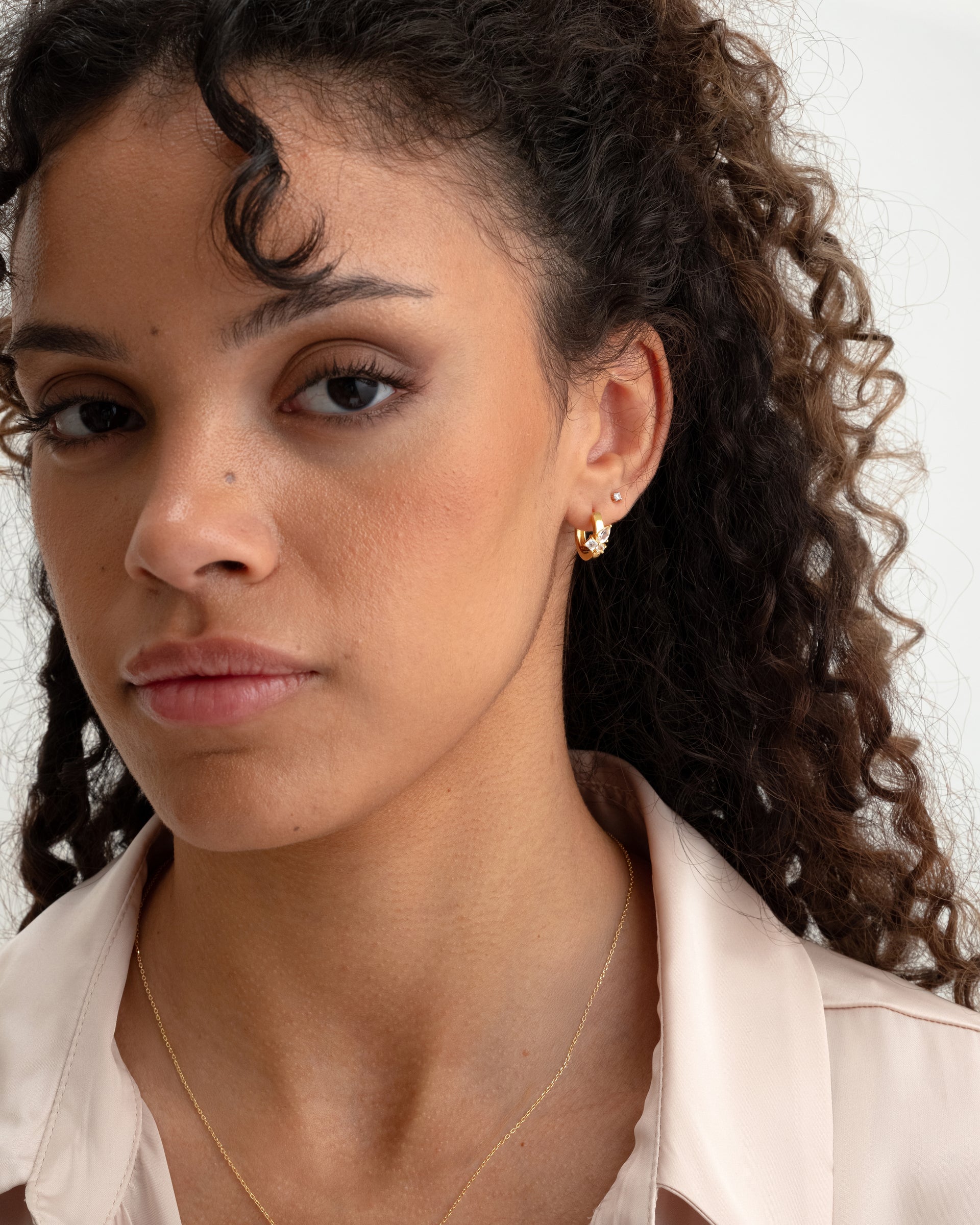 A close-up of a model wearing the gold Pam Hoops, adorned with white topazes, along with a small stud earring and a fine gold chain necklace.