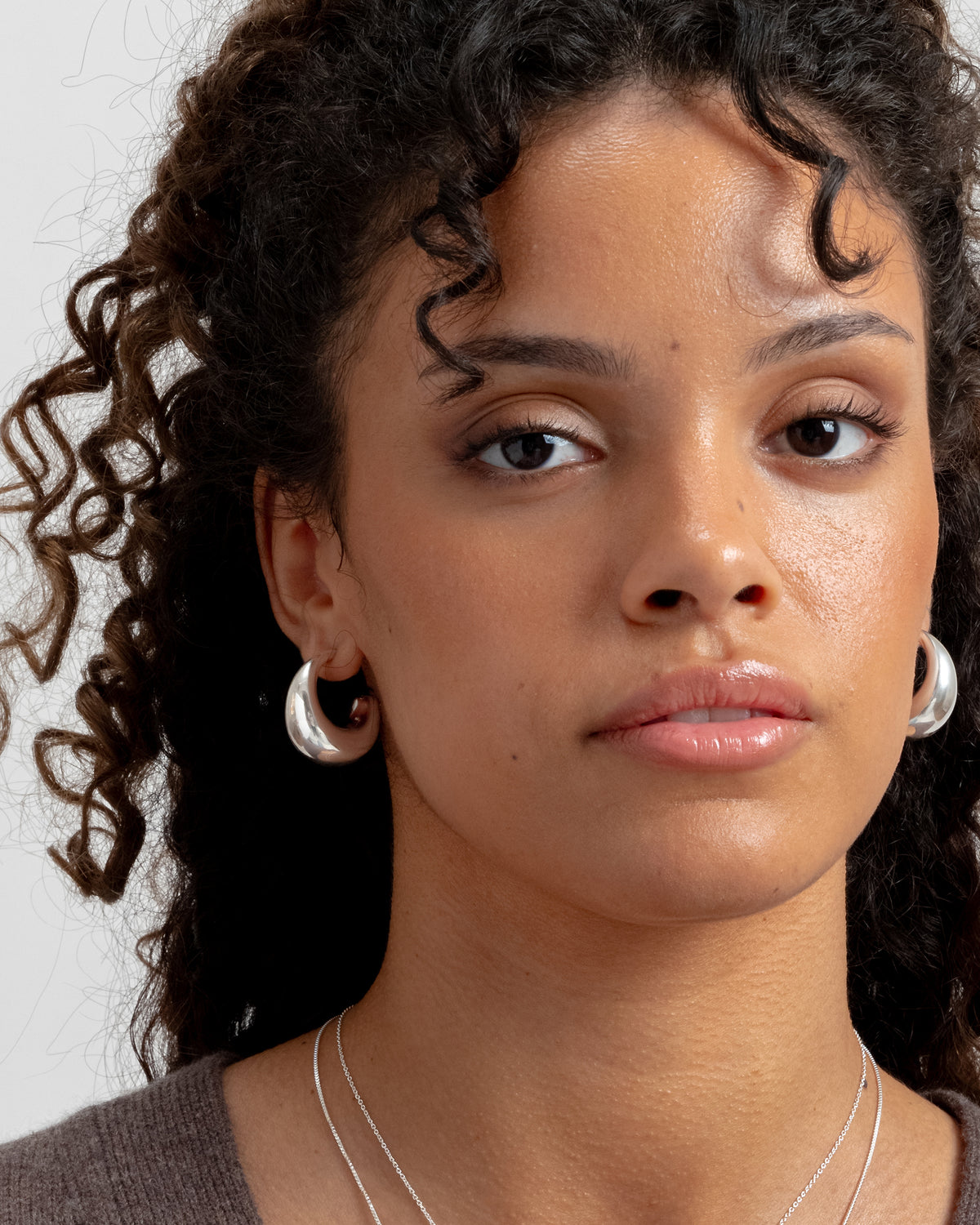 A close-up shot of a model wearing the Bolded Earrings in silver, which are thick, uniquely shaped hoops, along with fine silver chain necklaces.