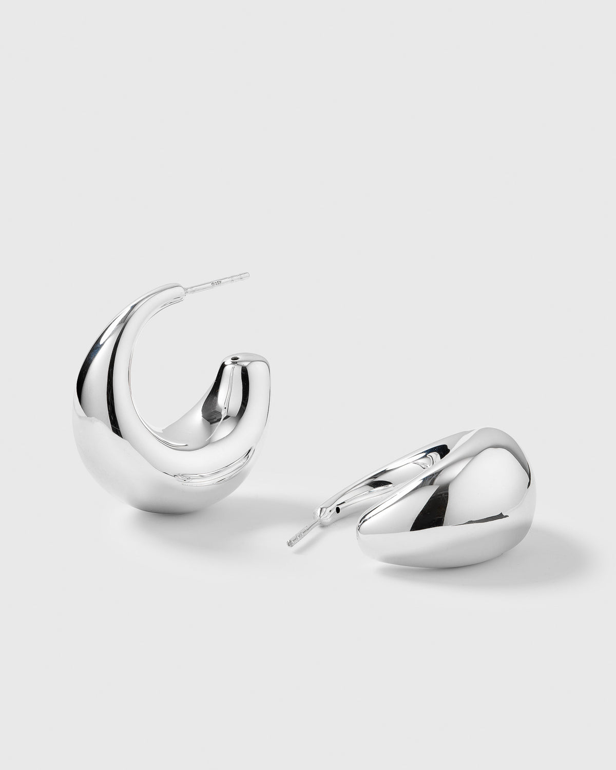 A product shot of the Bolded Earrings in silver, one standing and one lying down to display their unique, chunky, and curved hoop shape.