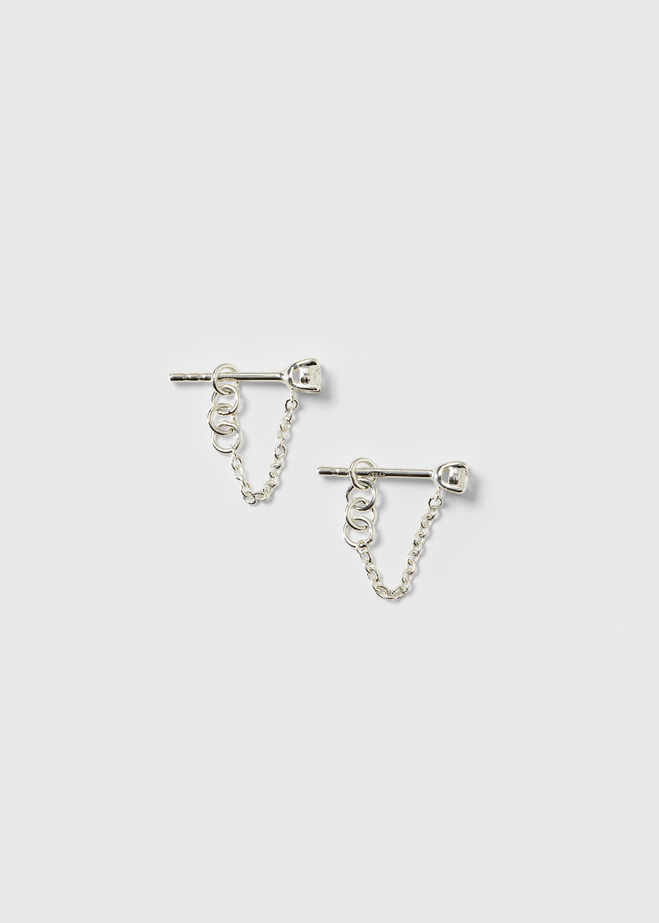 A product shot of the Minimalistica Solo Nova Earrings in silver, featuring a stud design with a white topaz and a delicate dangling chain.