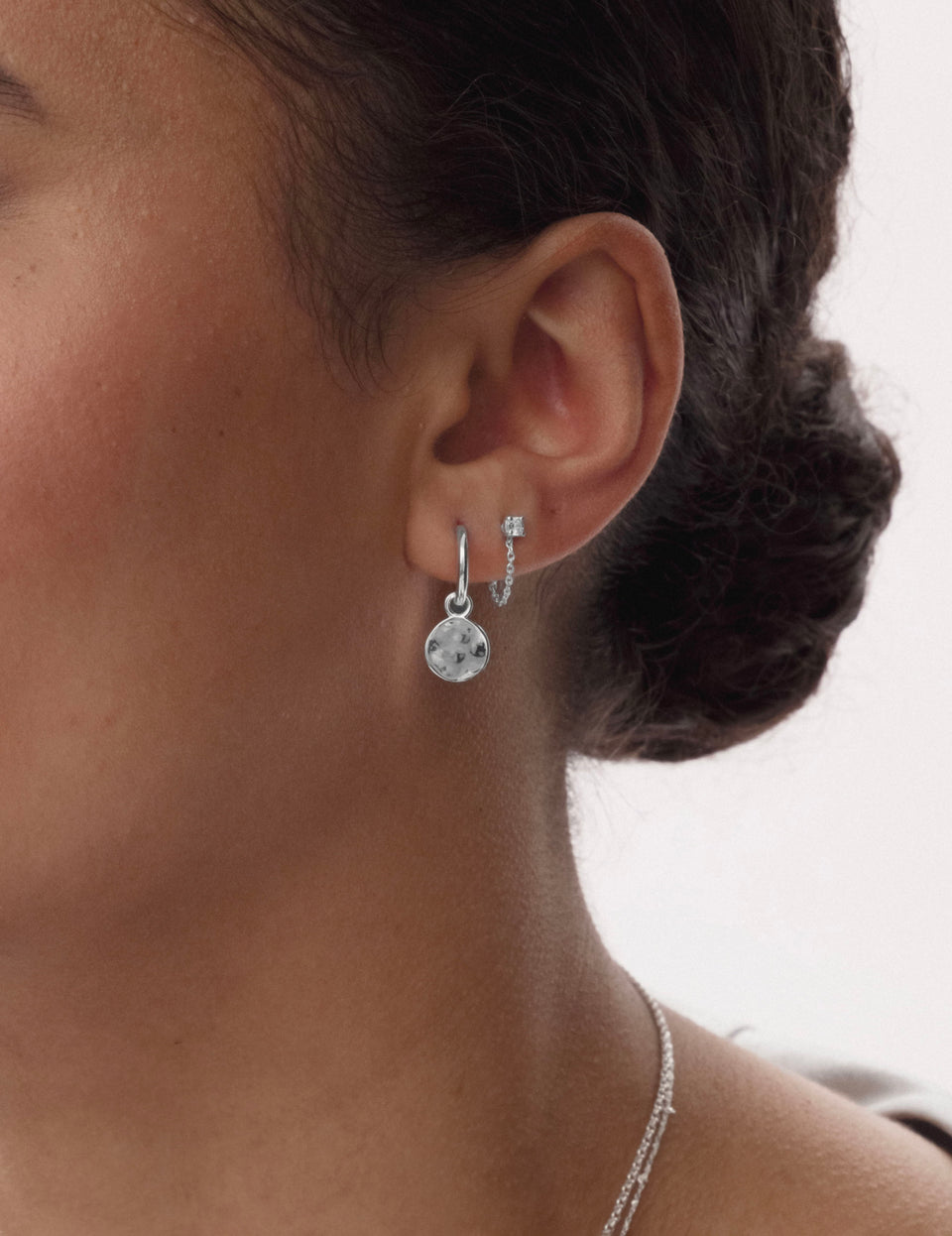A close-up of a model wearing the Minimalistica Solo Nova Earrings in silver, featuring a white topaz stud with a chain, paired with a silver hoop earring with a round hammered charm, and a delicate silver necklace.