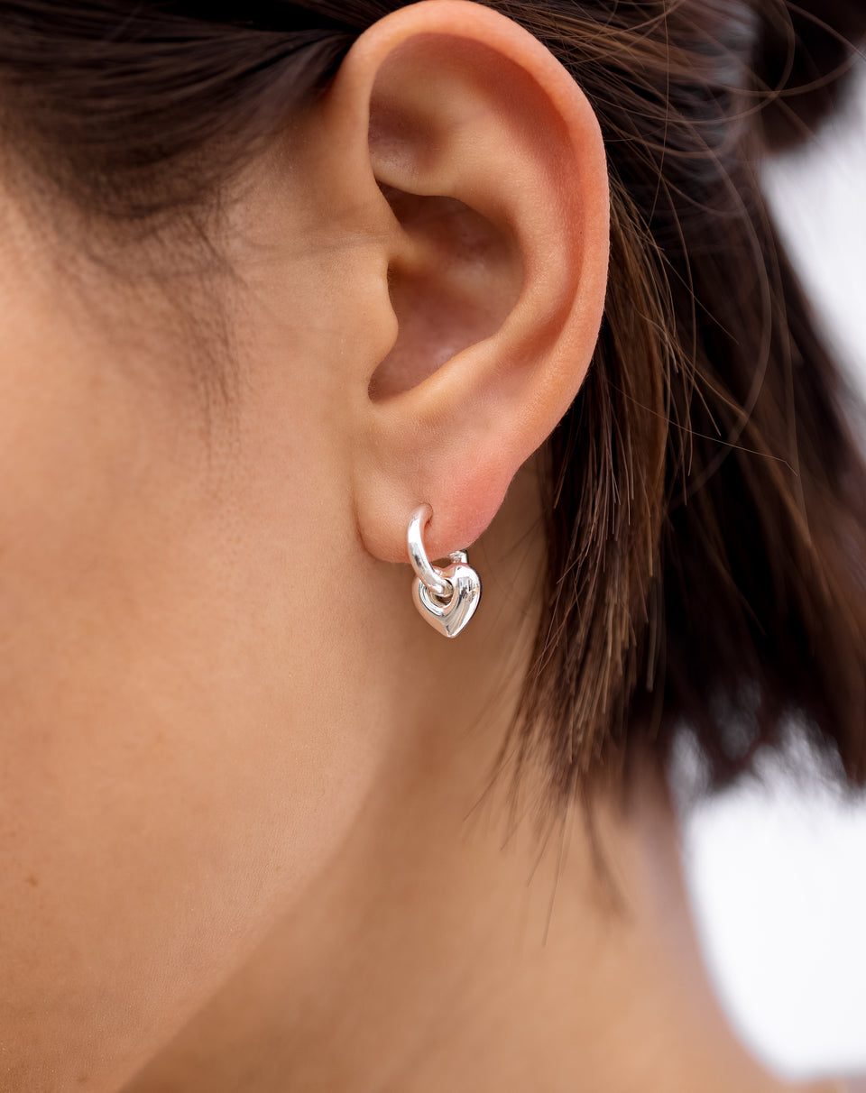 Close-up of a model's ear wearing the La La Love Fat Heart Earring in silver, a small hoop with a dangling, puffy heart-shaped charm.