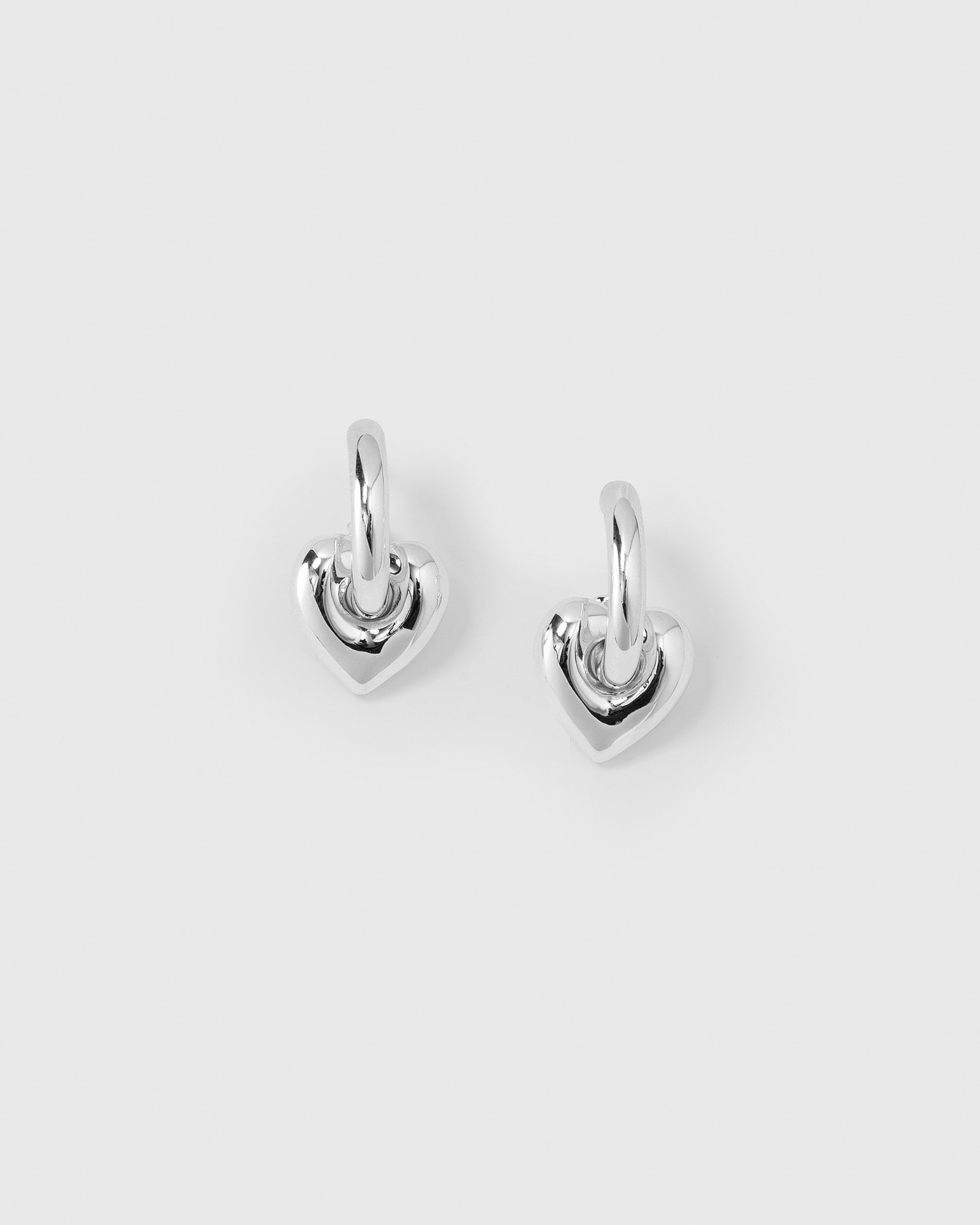 A product shot of the La La Love Fat Heart Earrings in silver. The image displays a pair of small hoops, each featuring a chunky, dangling heart charm.