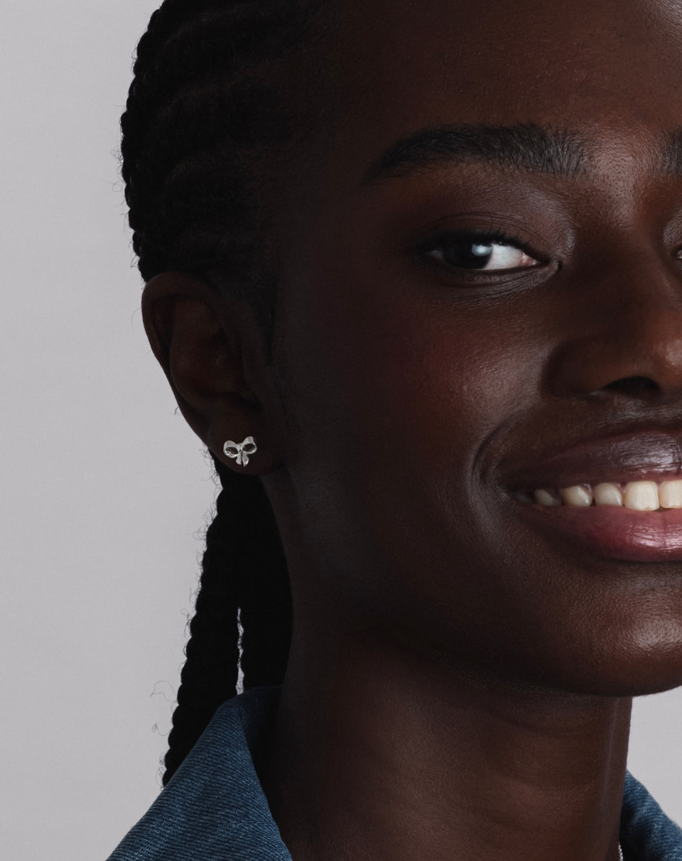 A close-up shot of a model wearing the Rosie Mini Studs Silver, a delicate stud earring shaped like a bow.