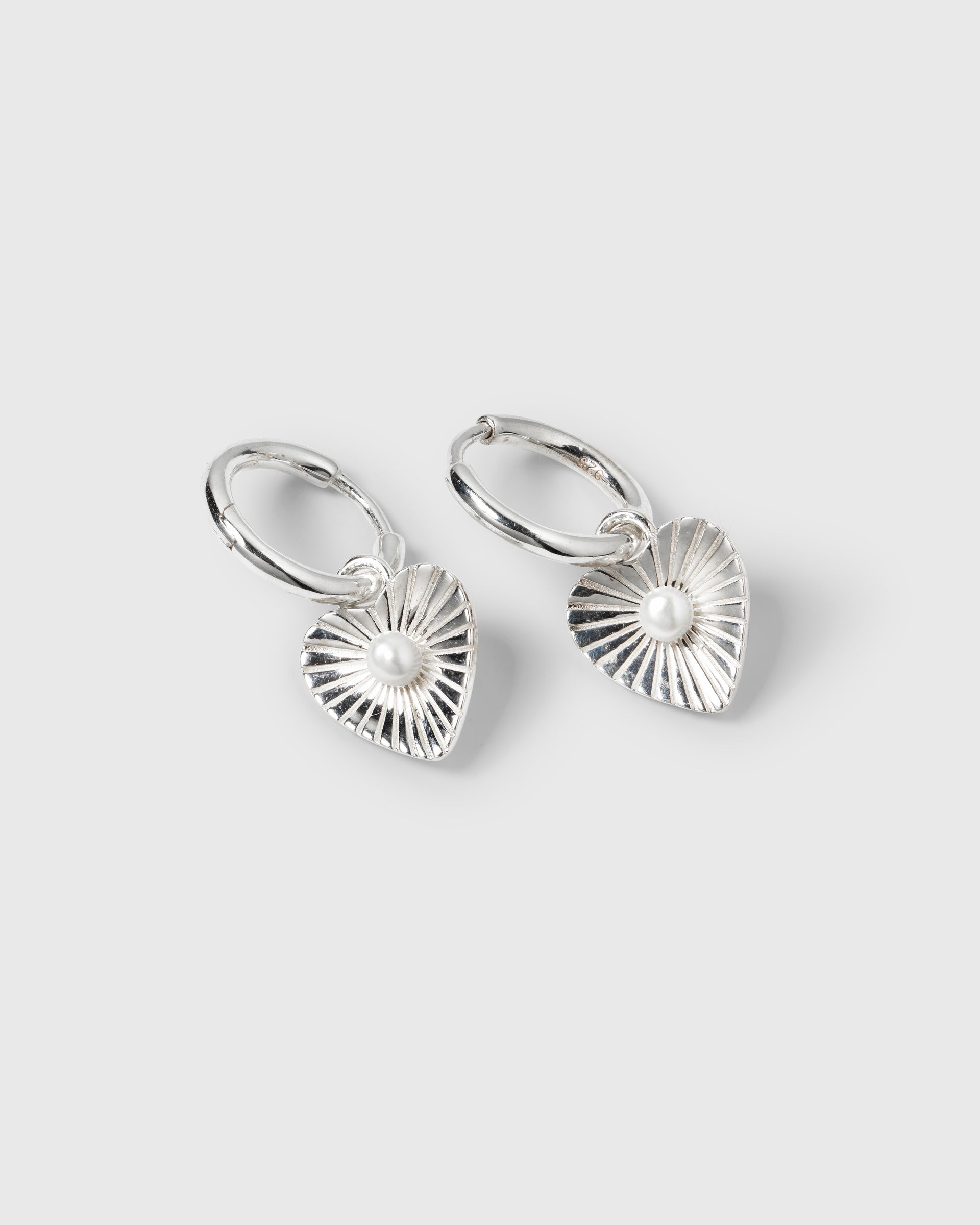 A product shot of the Aurelia Earrings in silver, featuring a pair of hoops with heart-shaped pendants, each detailed with a sunburst pattern and a central freshwater pearl.