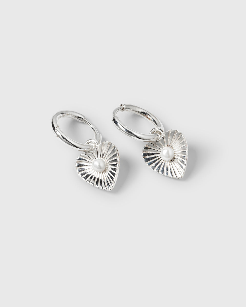 A product shot of the Aurelia Earrings in silver, featuring a pair of hoops with heart-shaped pendants, each detailed with a sunburst pattern and a central freshwater pearl.