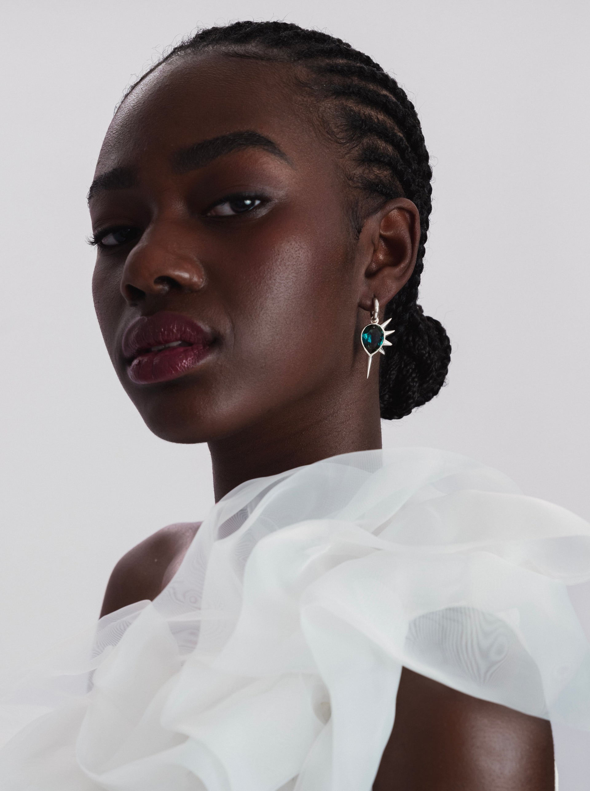 A close-up shot of a model wearing the Queen Of Spikes Earrings in silver, featuring a dangling pendant with a large, teardrop-shaped teal quartz and a spiky silver design.