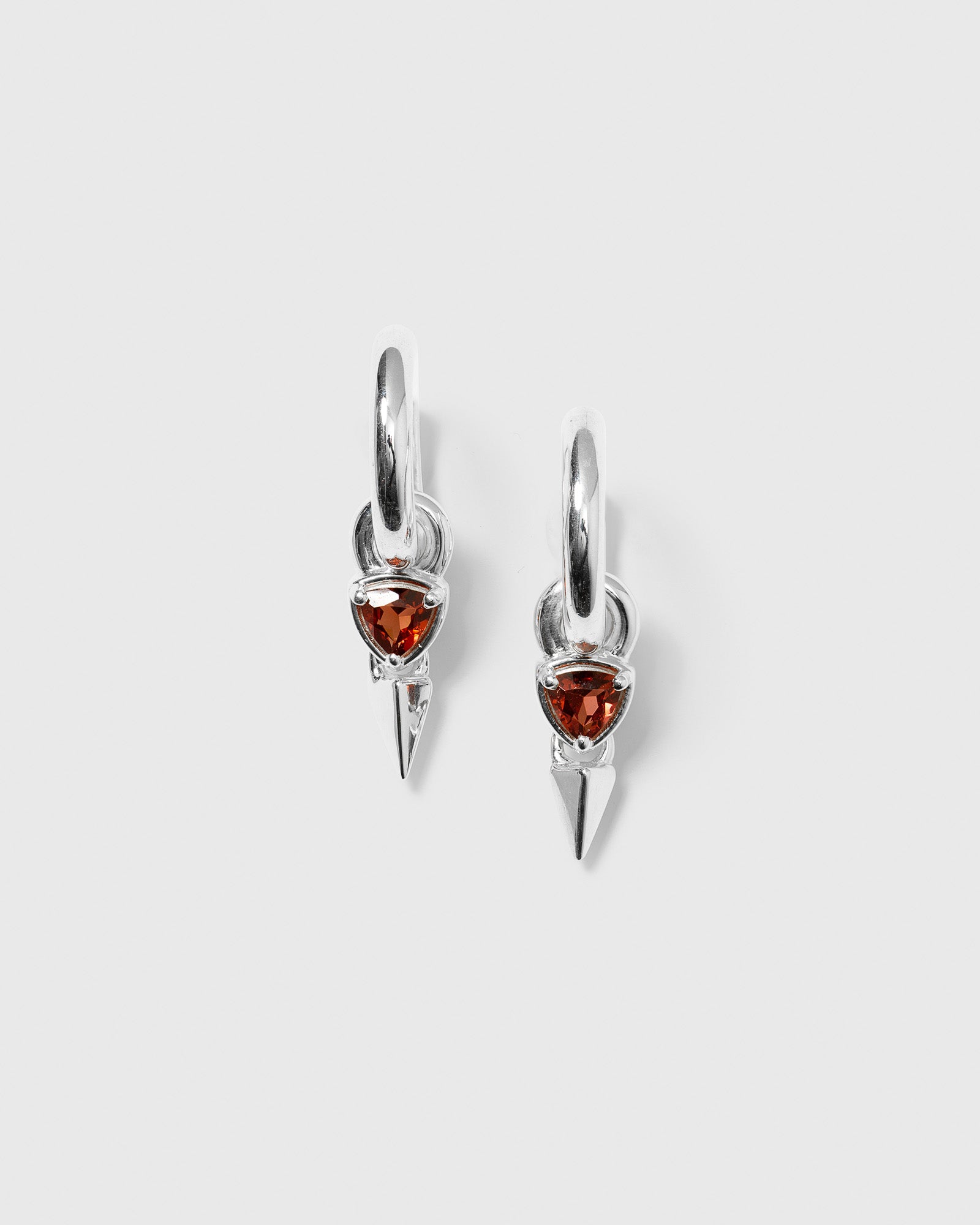 A product shot of the Mrs H Spike Earrings in silver, featuring chunky hoops with a dangling charm that has a triangular red garnet stone and a silver spike.