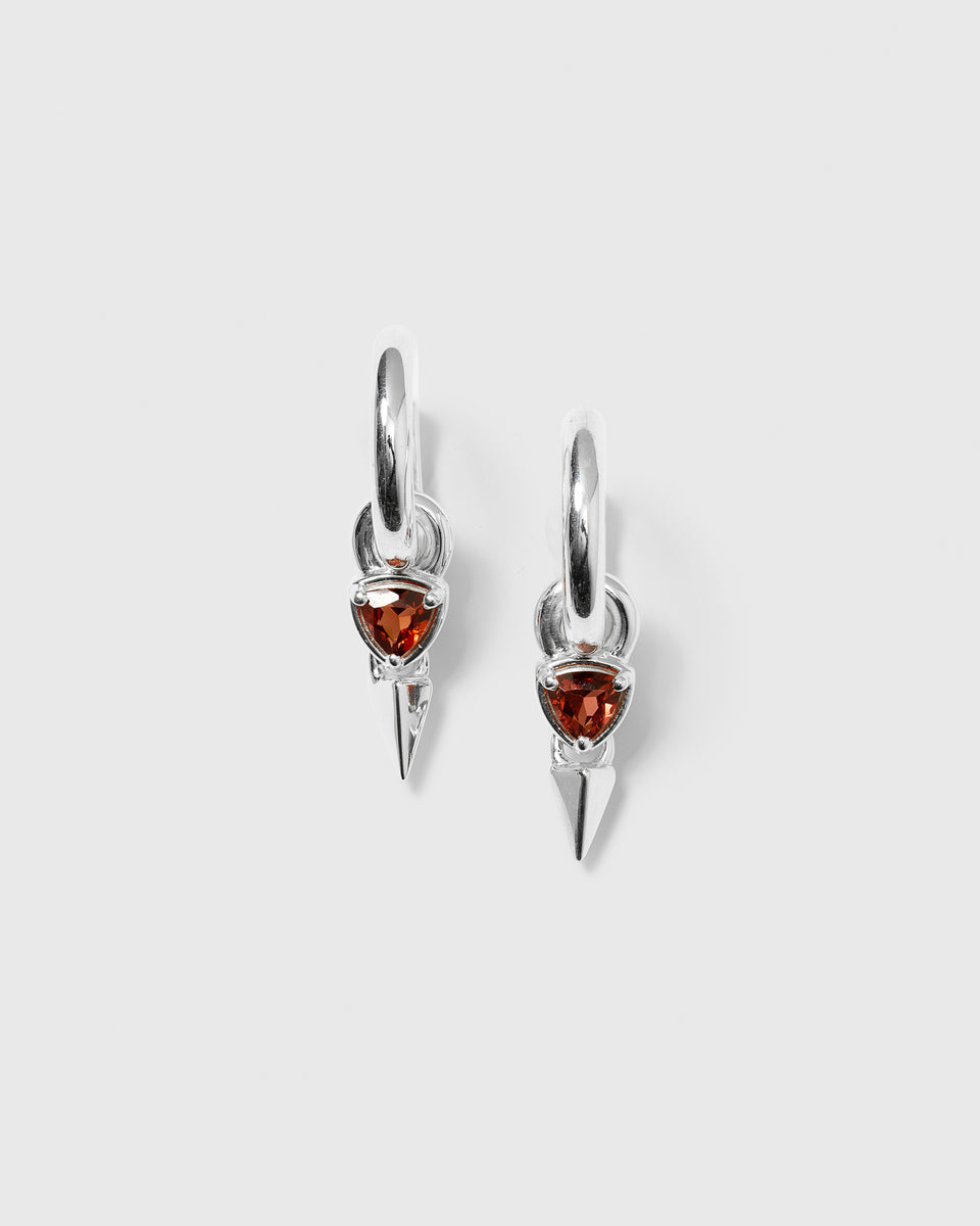 A product shot of the Mrs H Spike Earrings in silver, featuring chunky hoops with a dangling charm that has a triangular red garnet stone and a silver spike.