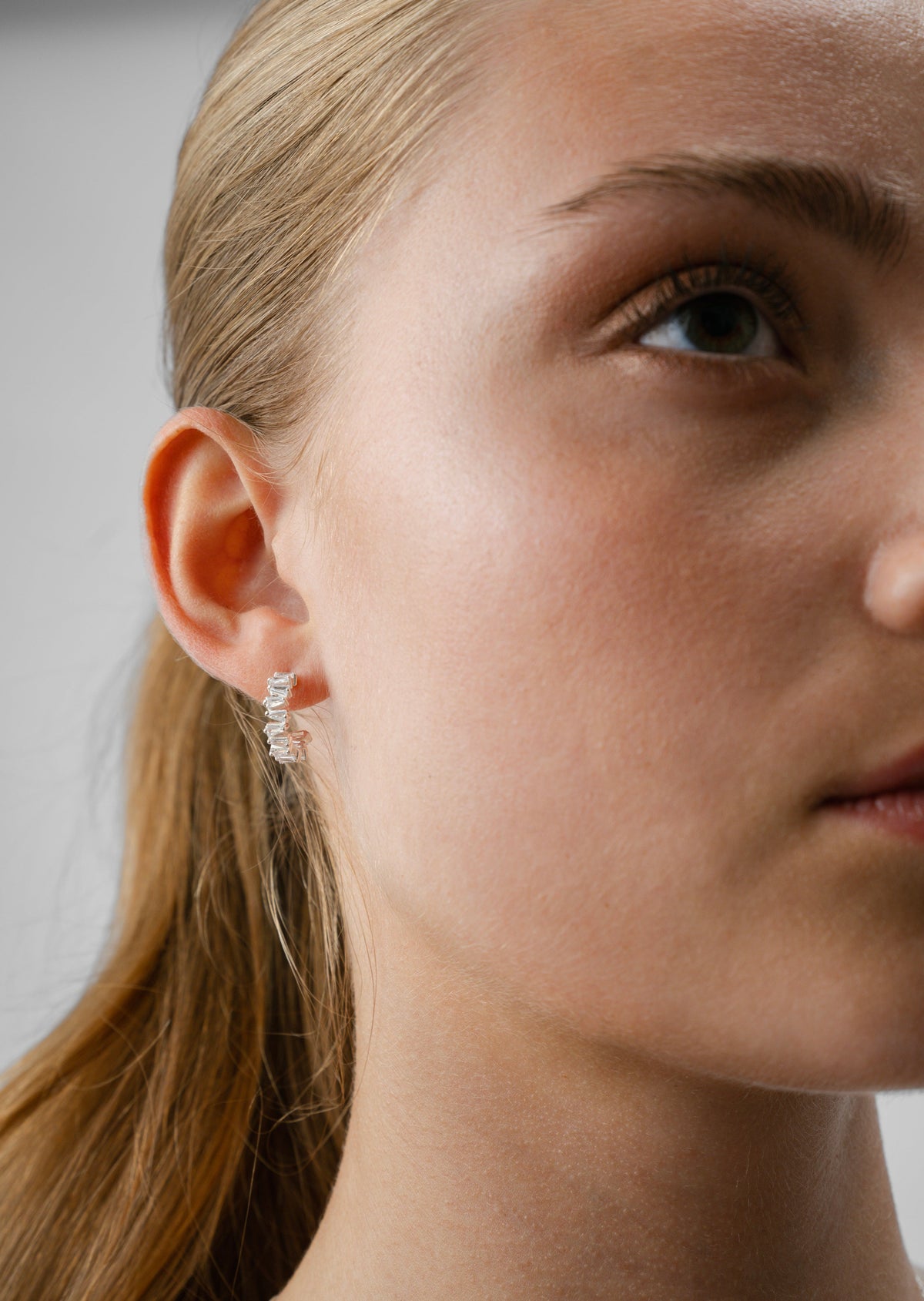 A close-up shot of a model wearing the Iggy Earrings in silver, small hoops adorned with sparkling, tapered baguette-cut cubic zirconias.