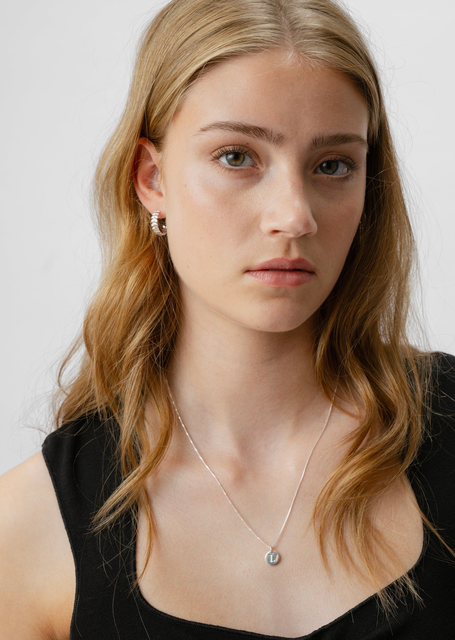 A model wears the Layers Olivia Hoops in silver, featuring a leaf-like pattern, paired with a simple silver chain necklace with a round 'L' initial pendant.