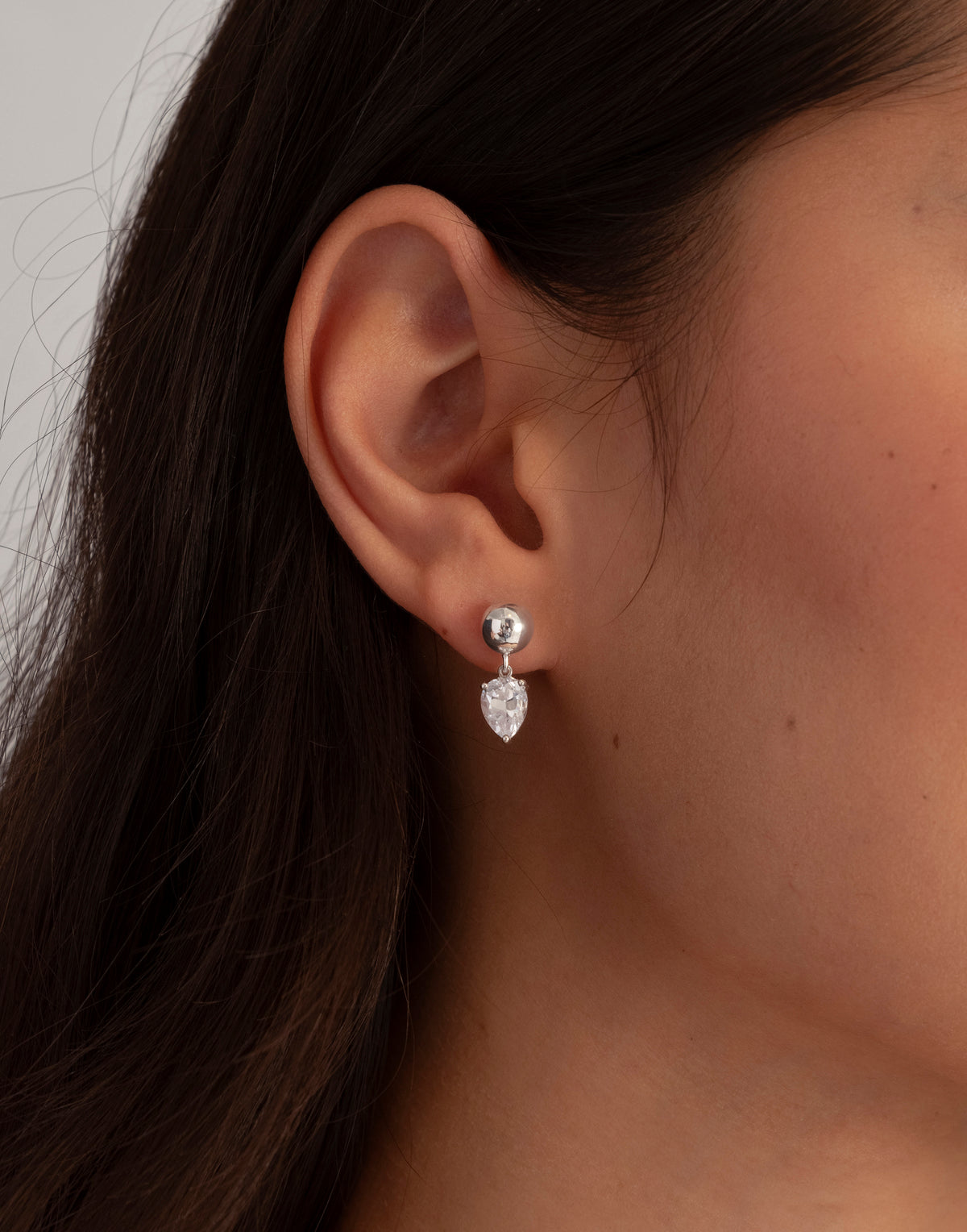 A close-up shot of a model wearing the Katie Earrings in silver, featuring a ball stud with a dangling drop-shaped cubic zirconia.