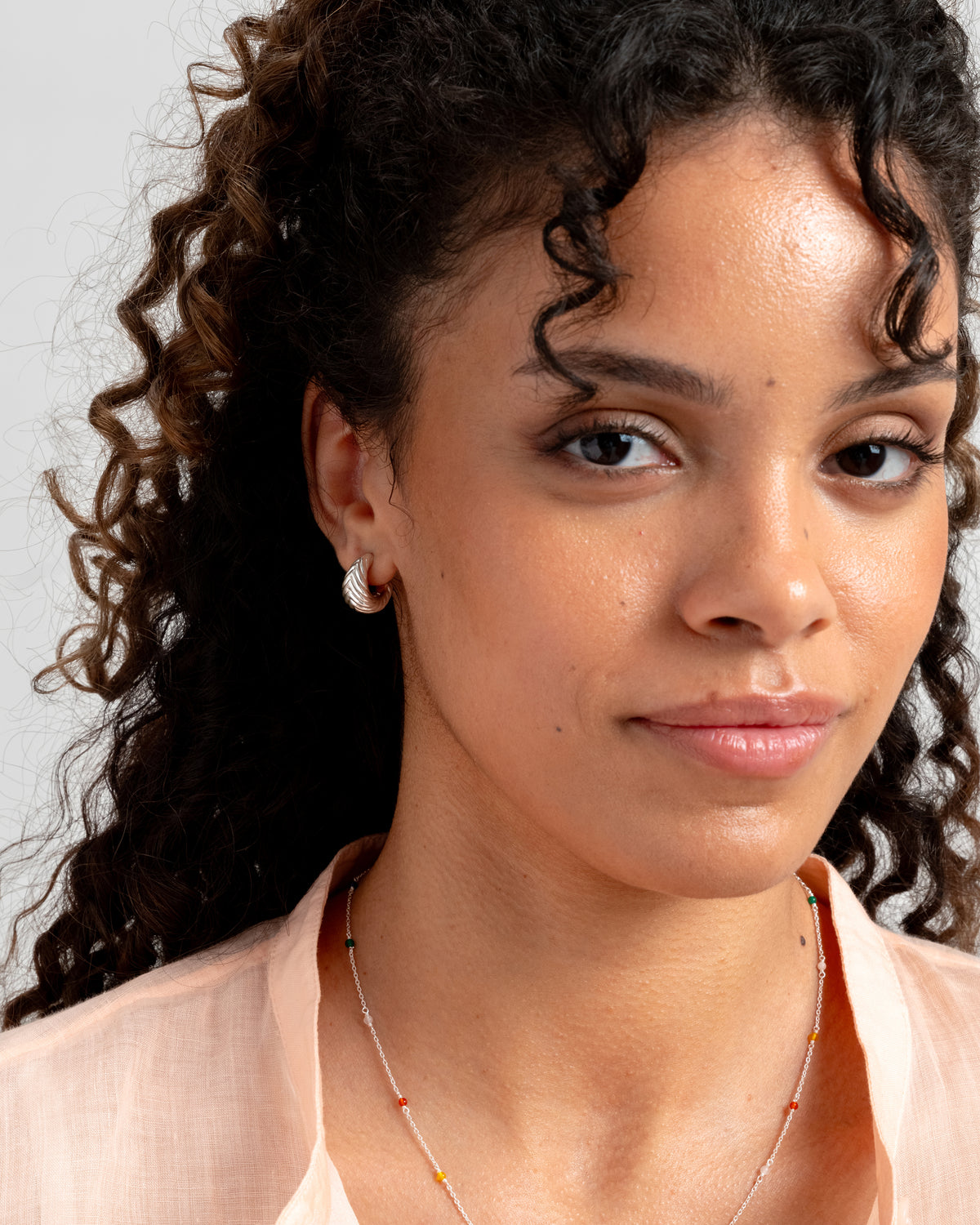 A close-up of a model wearing the Bolded Little Sis Chevron Earrings in silver and a delicate silver chain necklace with small, colorful beads.
