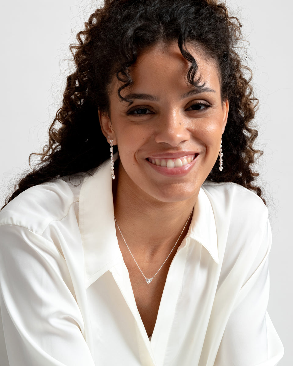 A smiling model wearing the Pam Long Earrings in silver, featuring a long, dangling design with sparkling white topaz stones, paired with a delicate silver necklace.
