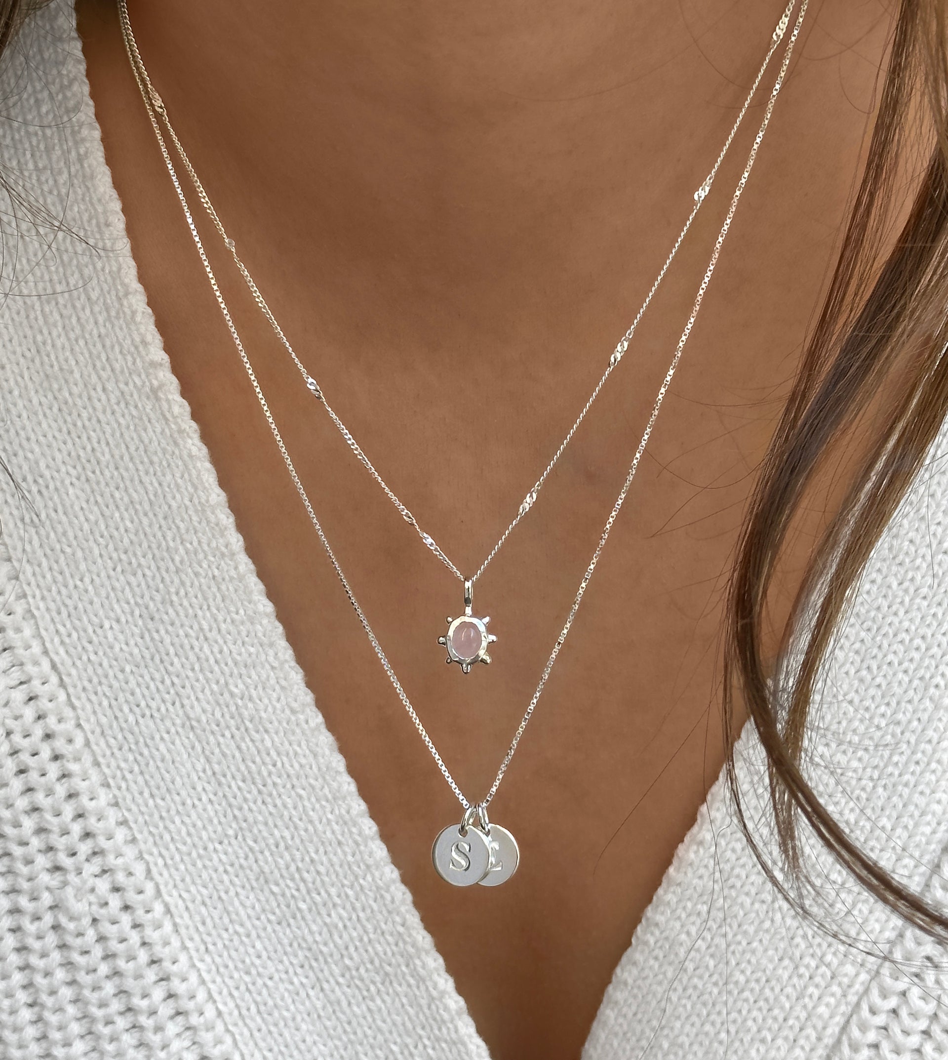 A close-up shot of a model wearing the Syster P Florentina Necklace in silver, layered with a second silver necklace. The Florentina features a flower-shaped pendant with a rose quartz stone on a delicate twisted chain, while the lower necklace has two round initial pendants.