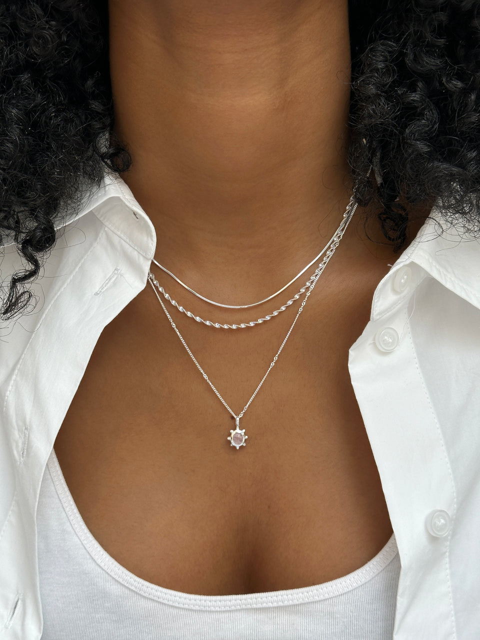 A close-up of a model wearing three layered silver necklaces from Syster P: a flat snake chain, a twisted rope chain, and the Florentina necklace featuring a flower-shaped pendant with a rose quartz stone.