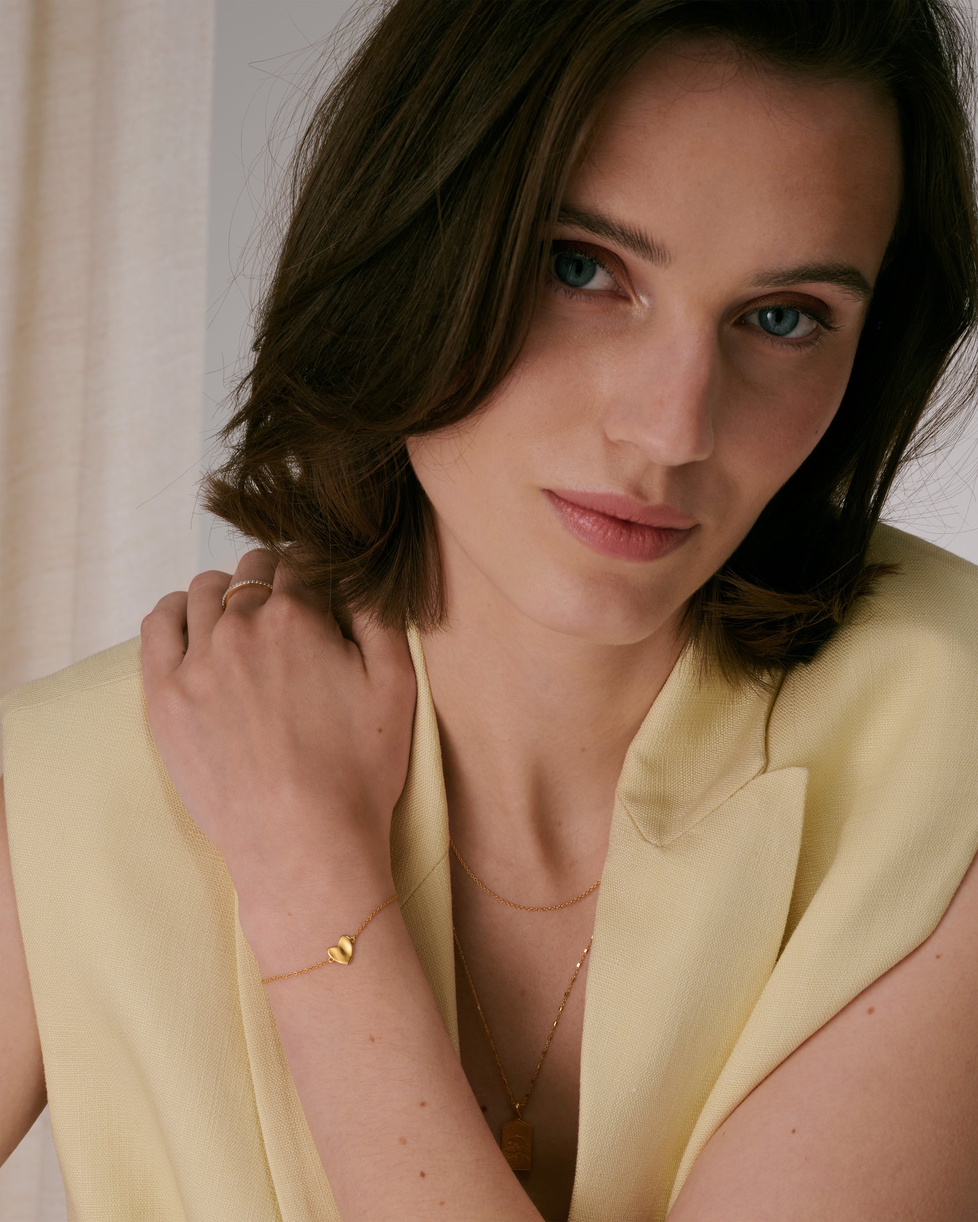 A delicate gold chain bracelet with a hammered heart charm, styled with a pave band ring and two layered gold necklaces.