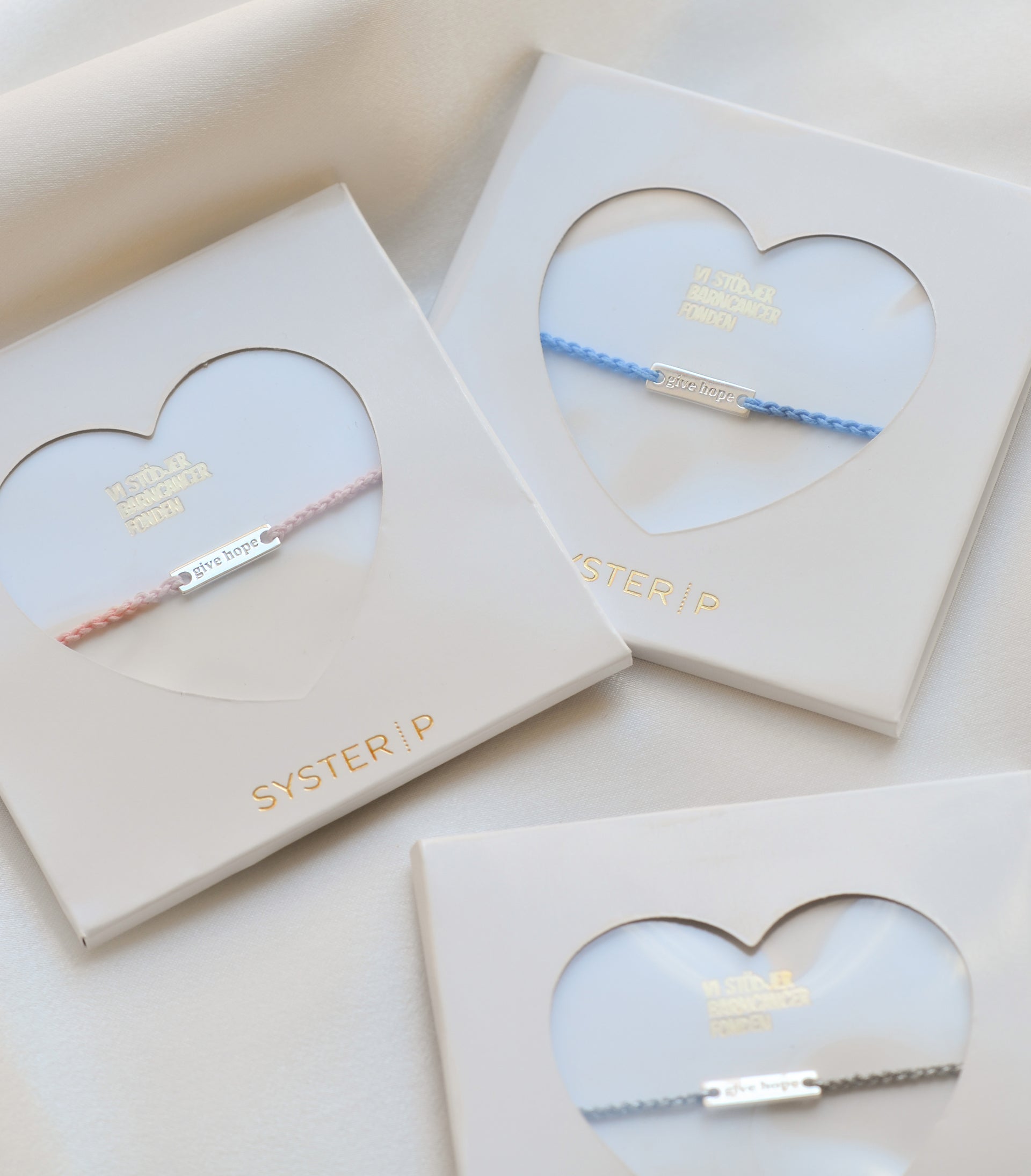 A product shot of the Give Hope thread bracelets in coral, blue, and grey, each presented in a white Syster P gift box with a heart-shaped window.