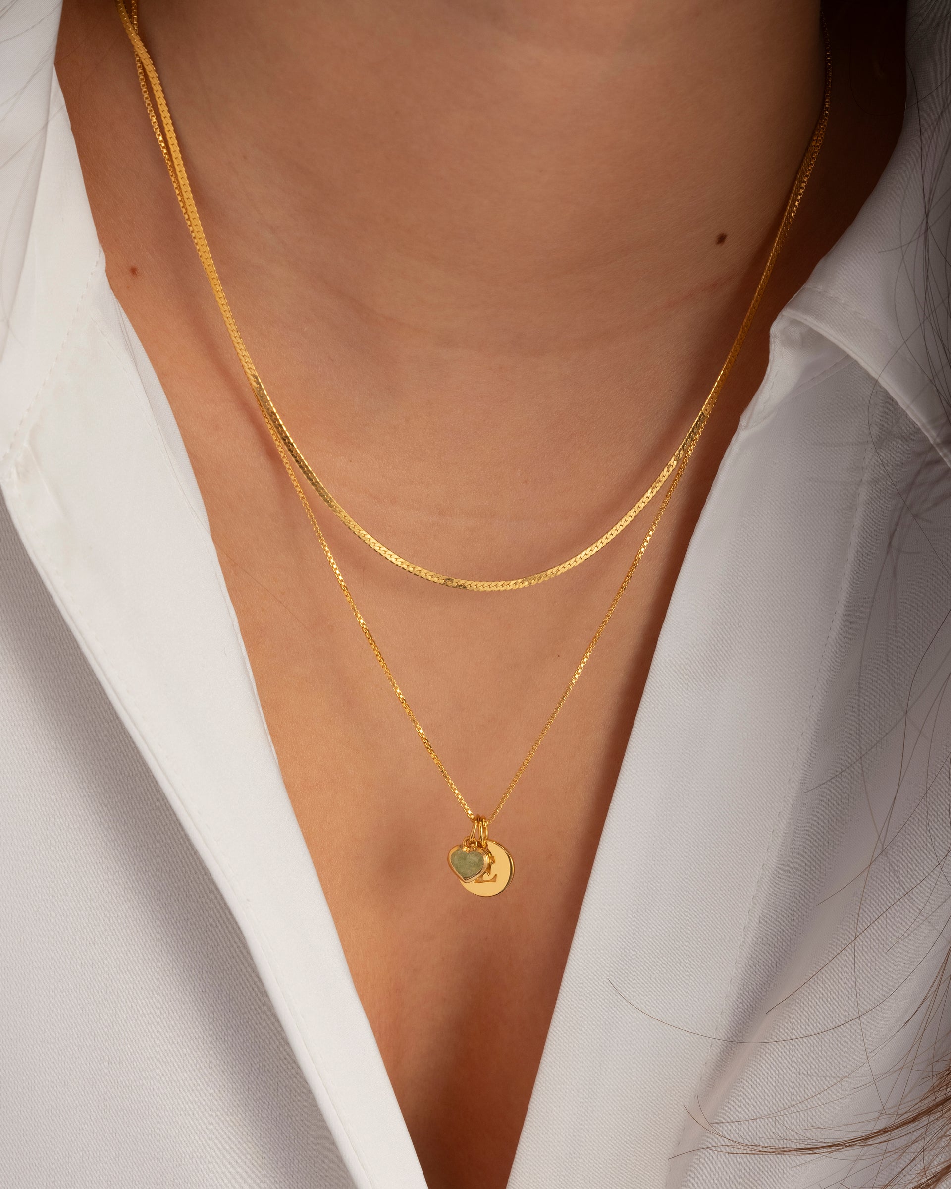 A close-up of a model wearing two layered gold necklaces; a flat herringbone chain and a thinner chain with the Beloved Heart Pendant in green aventurine paired with a round gold disc charm.